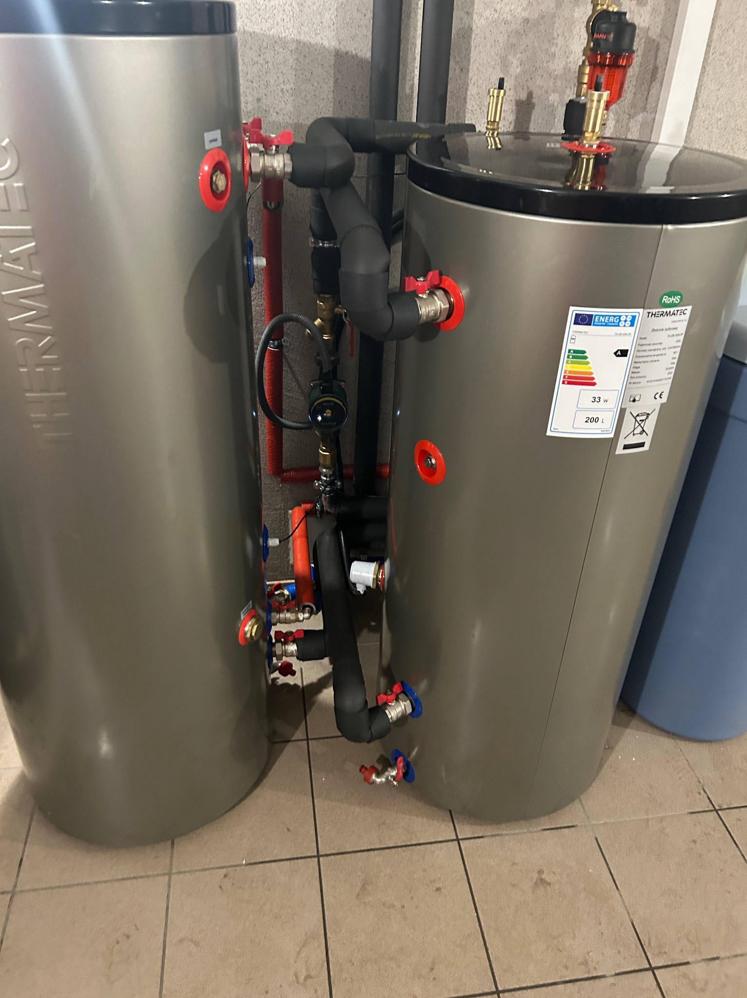 Dwa szare zbiorniki akumulacyjne Thermatec połączone rurami, z widocznymi zaworami i etykietą energetyczną na jednym z nich, na tle szarej ściany i podłogi z płytek.