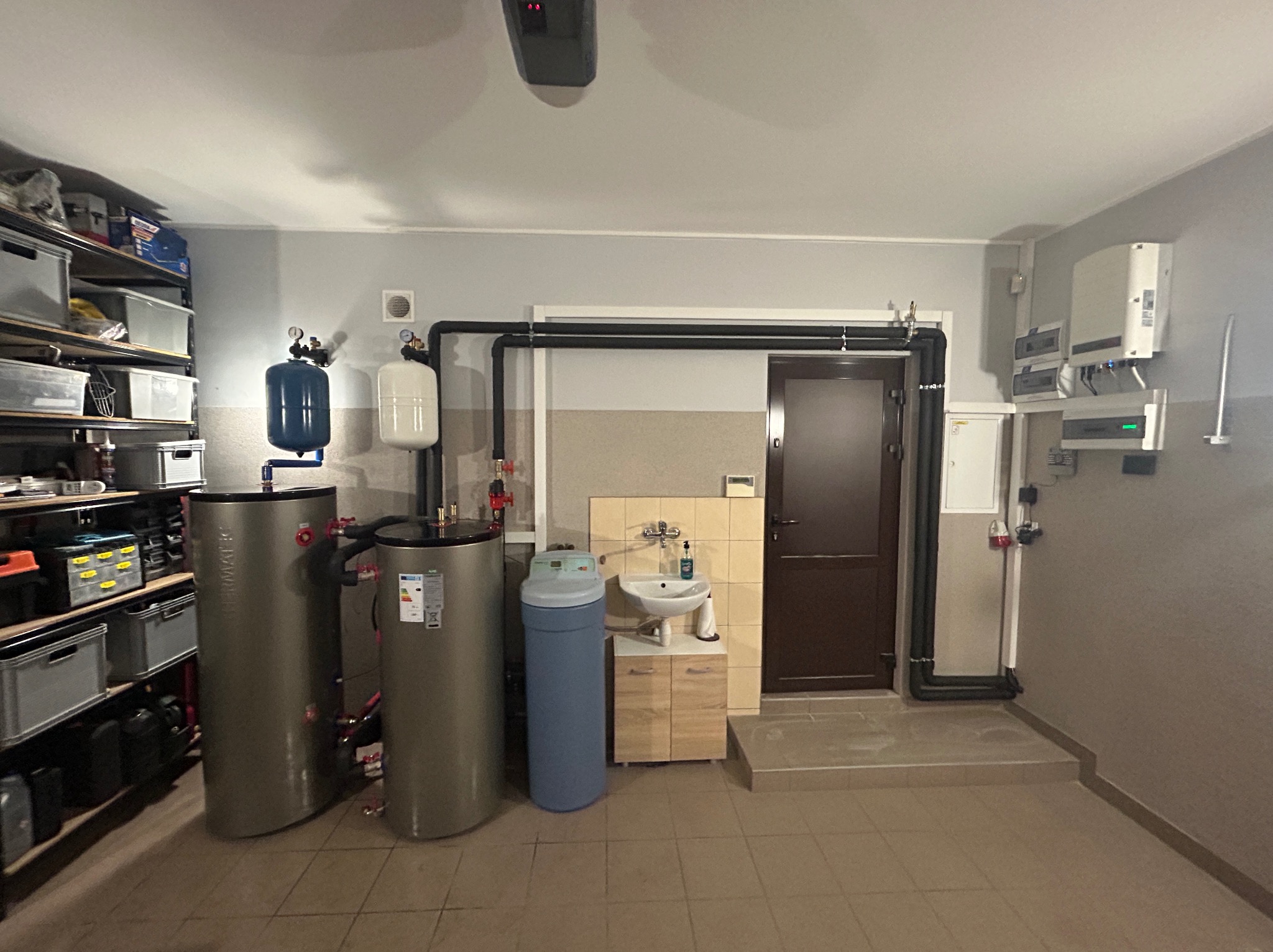 Dwa szare zbiorniki akumulacyjne ciepłej wody z niebieskim naczyniem przeponowym, obok zmiękczacz wody, całość w pomieszczeniu gospodarczym z regałem i instalacją fotowoltaiczną na ścianie.