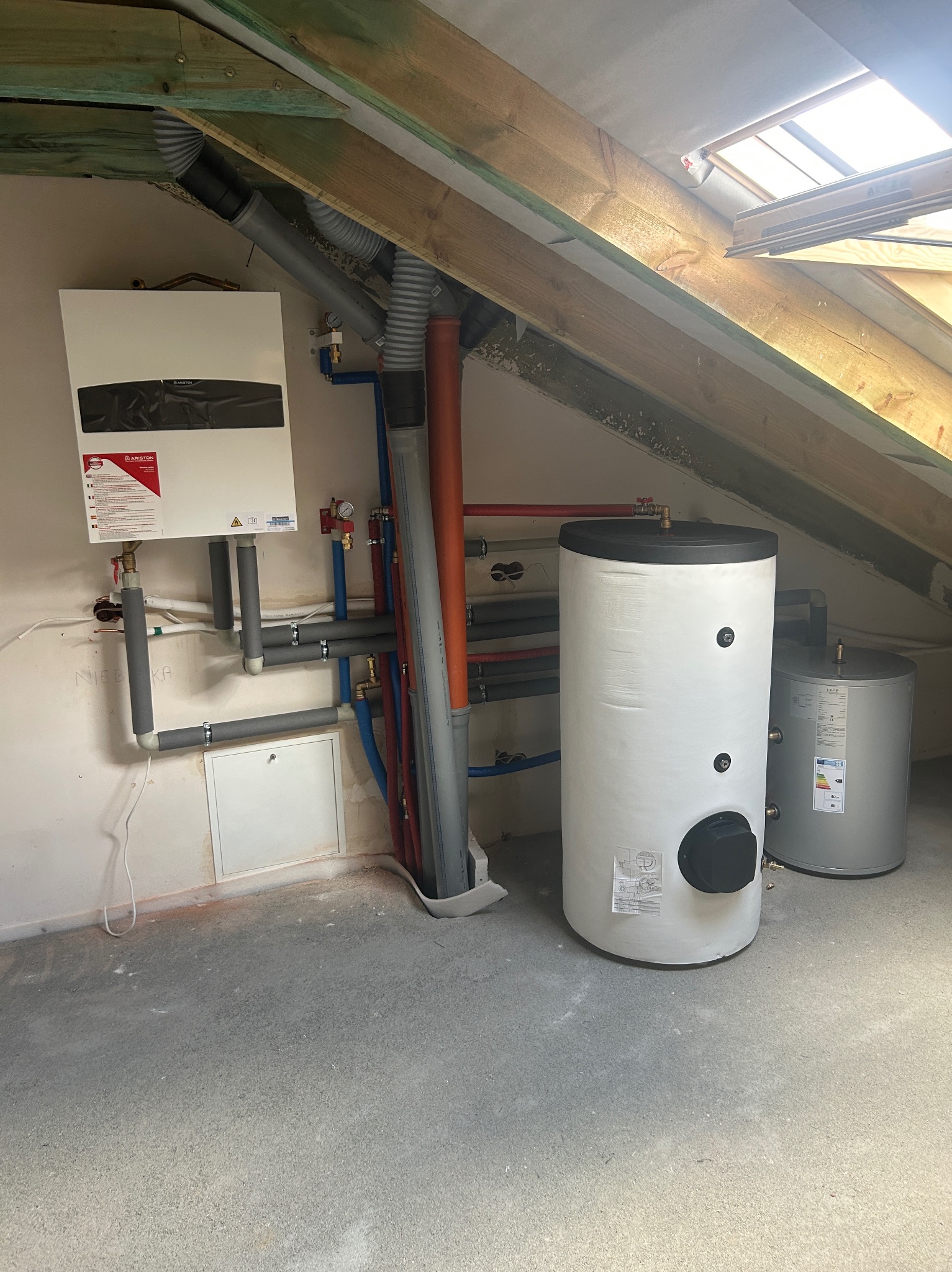 Biały piec gazowy Ariston z podłączonymi rurami w kotłowni na poddaszu, widoczny również biały bojler i szary zbiornik akumulacyjny Joule, drewniana konstrukcja dachu ze świetlikiem.