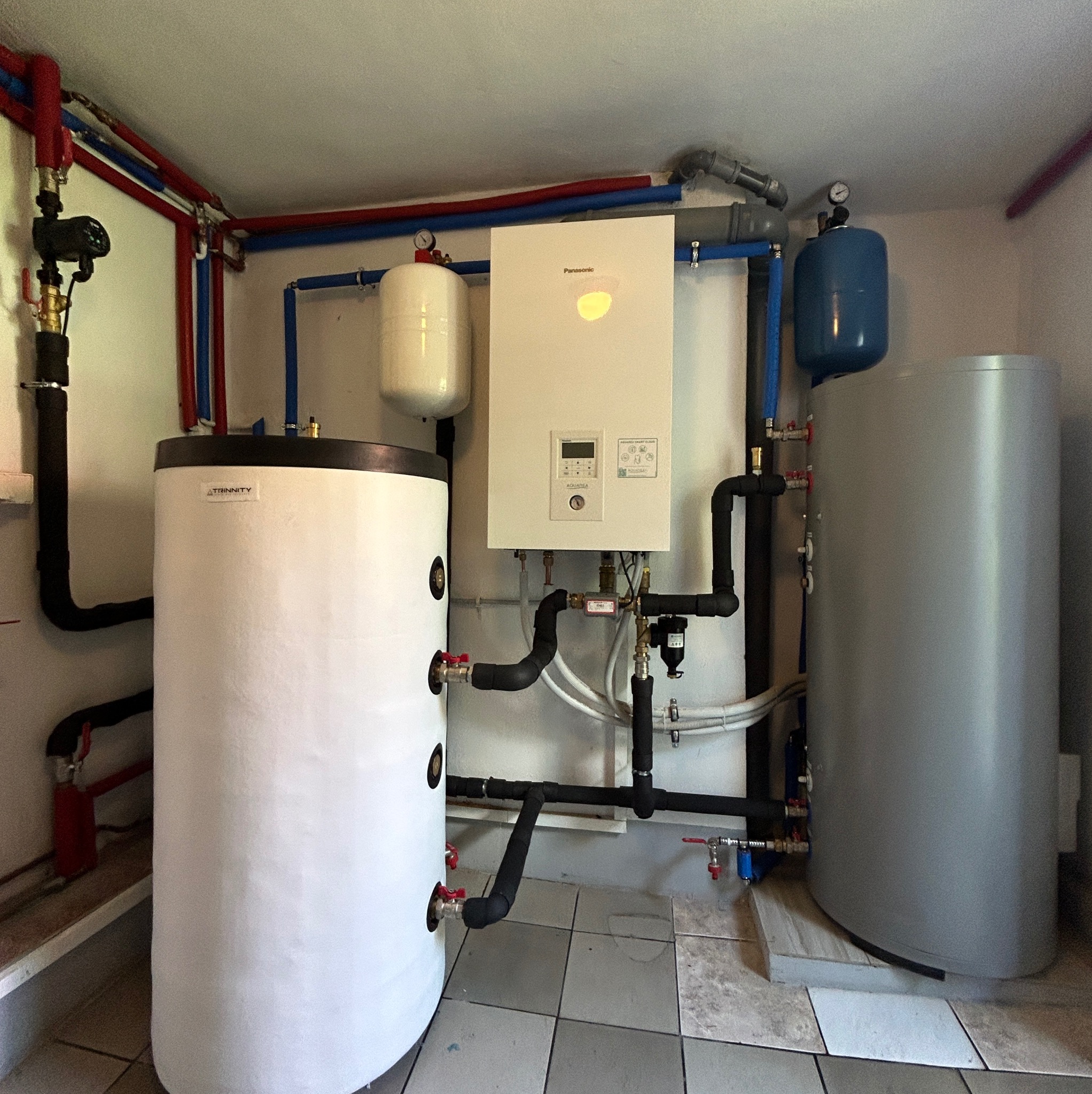 Kompaktowa kotłownia z białym piecem gazowym Panasonic, zbiornikami buforowymi i instalacją hydrauliczną z rur w kolorach czerwonym i niebieskim.