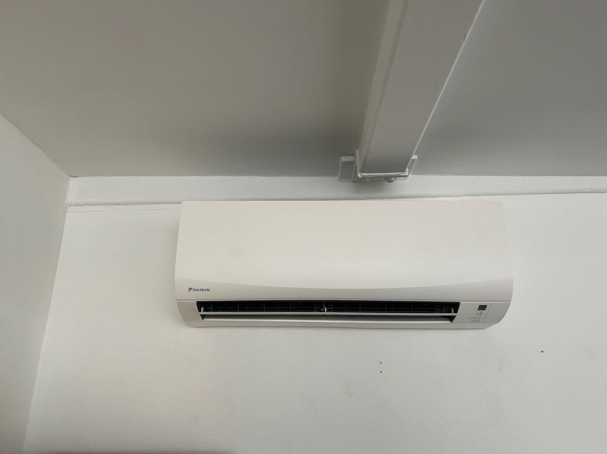 Biała jednostka wewnętrzna klimatyzatora marki Daikin zamontowana na białej ścianie, widoczny fragment białej rury pod sufitem.