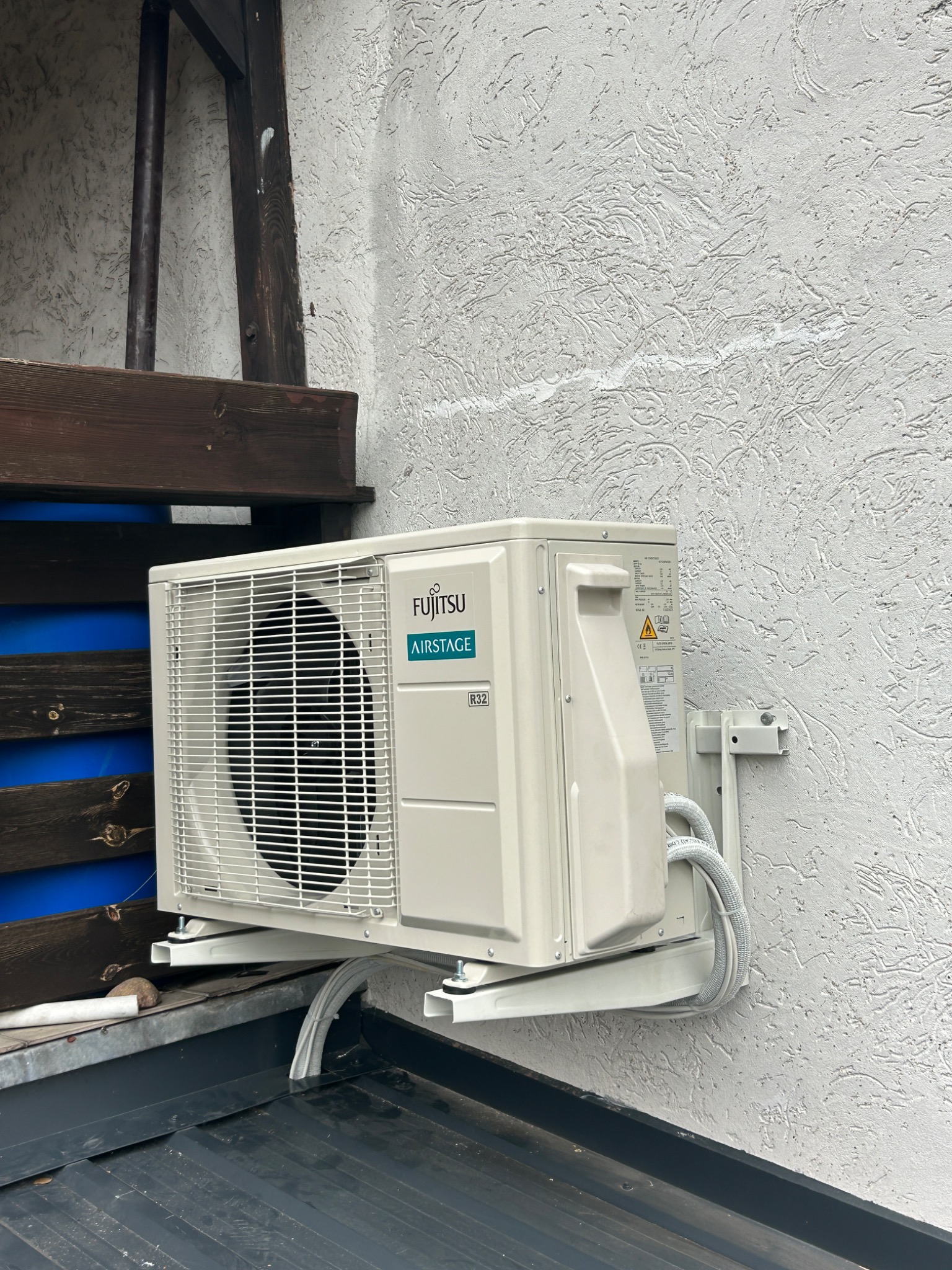 Zewnętrzna jednostka klimatyzacji Fujitsu Airstage zamontowana na wspornikach przy teksturowanej, jasnej ścianie budynku; widoczne przewody w peszlach i fragment dachu pokrytego blachą na rąbek...