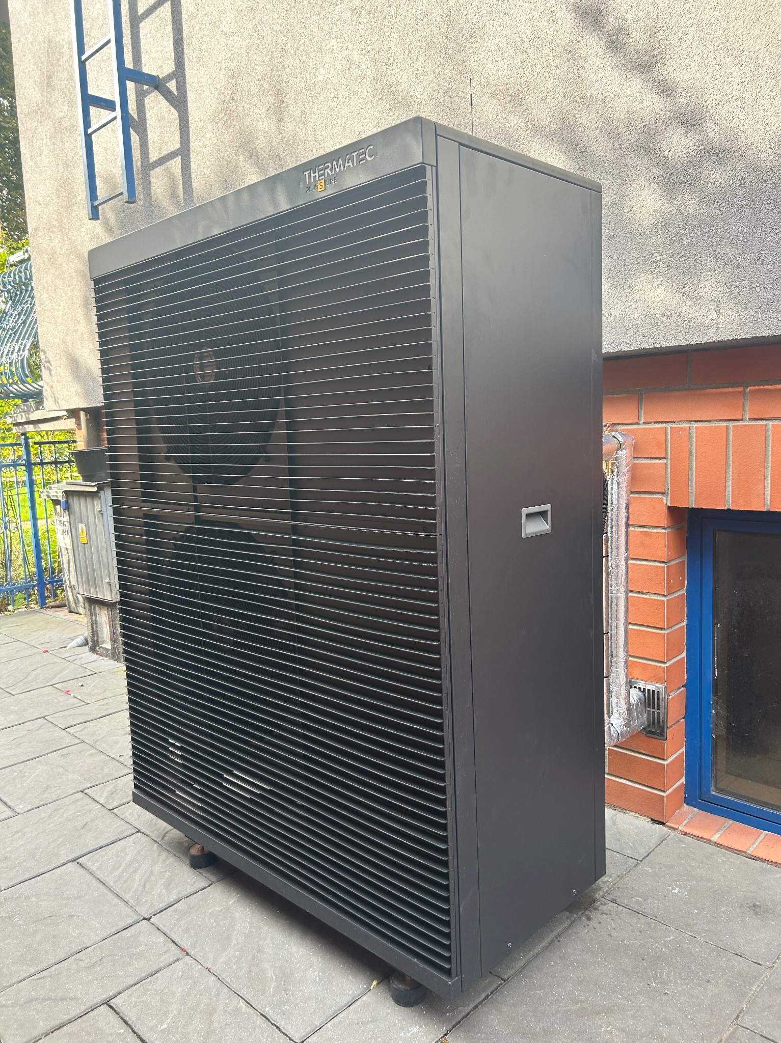 Czarna pompa ciepła Thermatec Plus S Line ustawiona na szarych płytach chodnikowych przy ścianie budynku z cegły i oknem z niebieską ramą. Widoczna rura odprowadzająca skropliny.