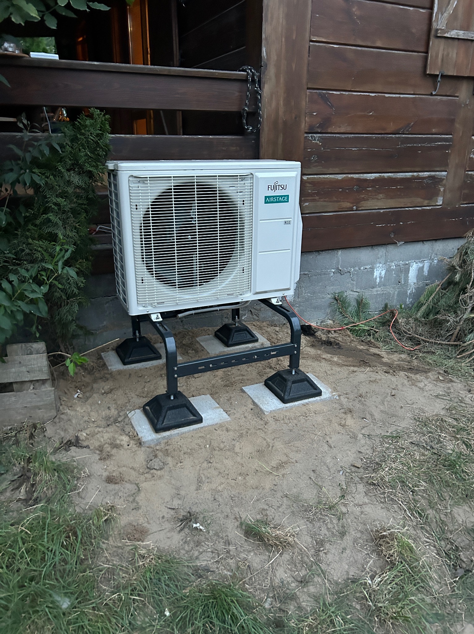 Biała jednostka zewnętrzna klimatyzatora Fujitsu Airstage, zamontowana na czarnej metalowej ramie z betonowymi podkładkami, stoi przy drewnianej ścianie domu.