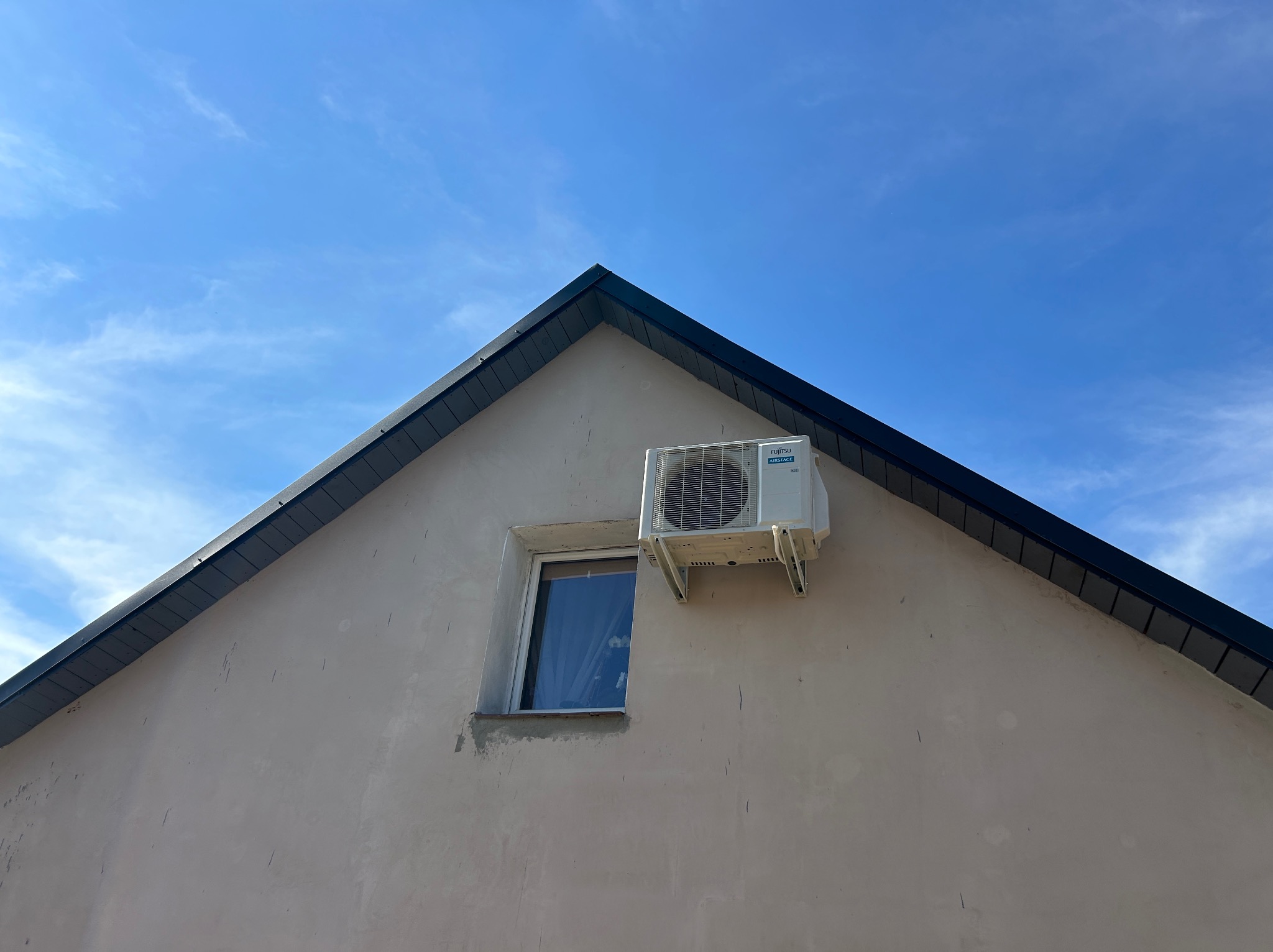 Biała jednostka zewnętrzna klimatyzatora marki Fujitsu zamontowana na elewacji domu obok okna, na tle błękitnego nieba z delikatnymi chmurami.