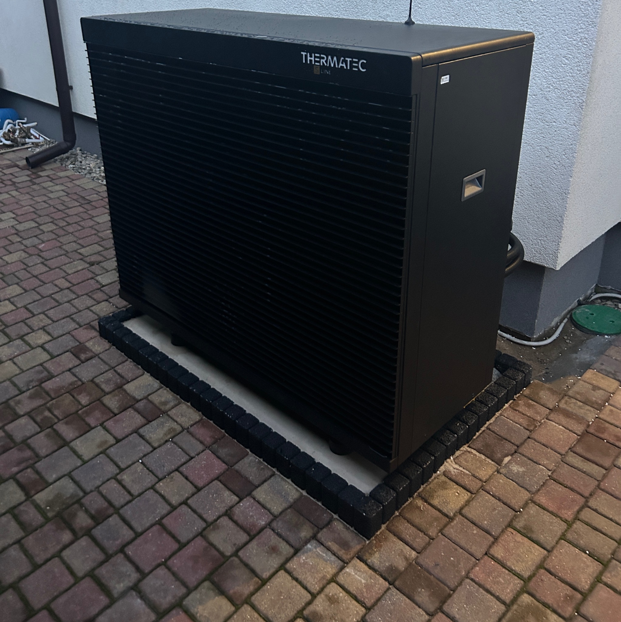 Czarna pompa ciepła Thermatec Line zainstalowana na kostce brukowej, widoczne przyłącza i fragment ściany budynku w tle.