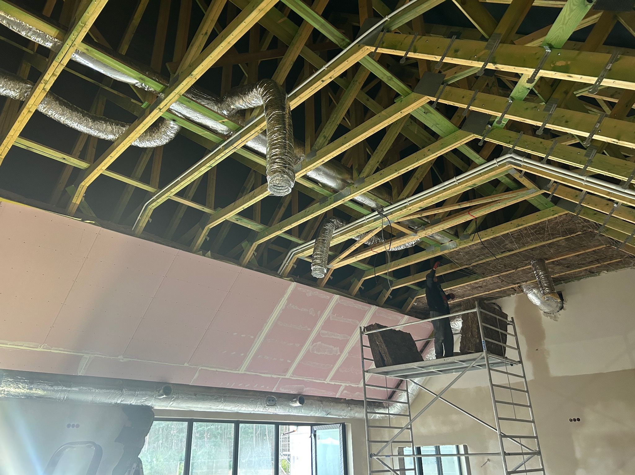 Instalacja przewodów wentylacyjnych w budynku z drewnianą konstrukcją dachu, widoczna osoba na rusztowaniu montująca izolację, różowe płyty kartonowo-gipsowe częściowo zamontowane na suficie.