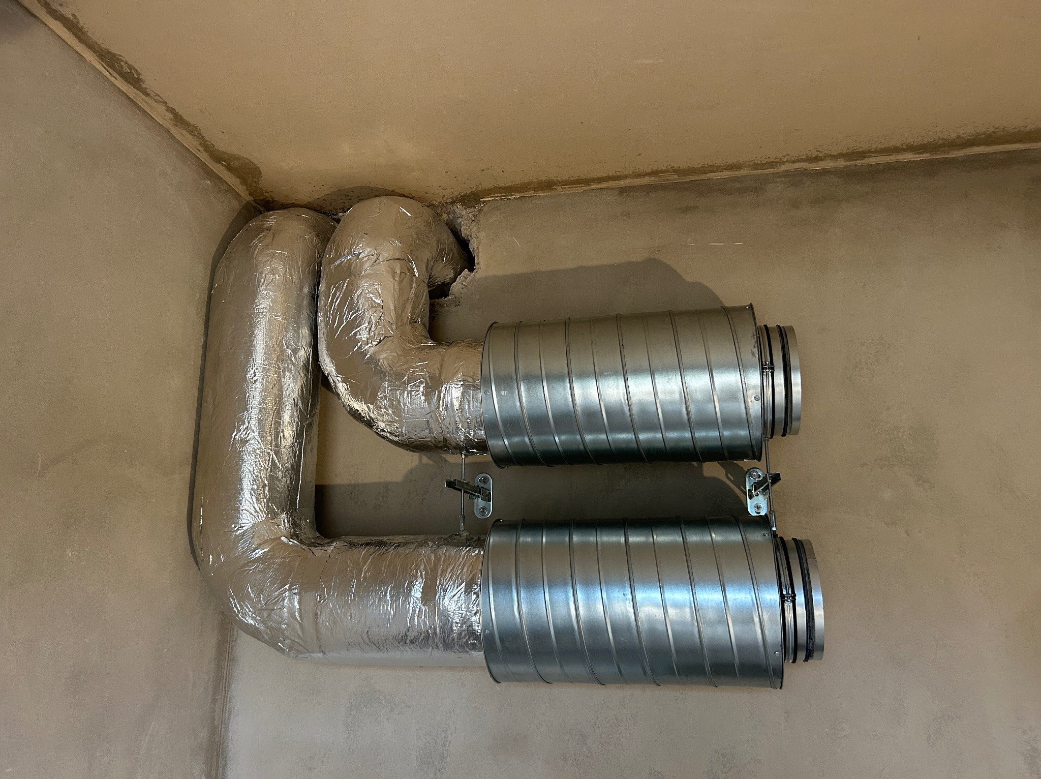 Dwa równoległe kanały wentylacyjne, jeden owinięty folią aluminiową, zamontowane na ścianie w surowym wykończeniu, z widocznymi śladami po kuciu w ścianie.