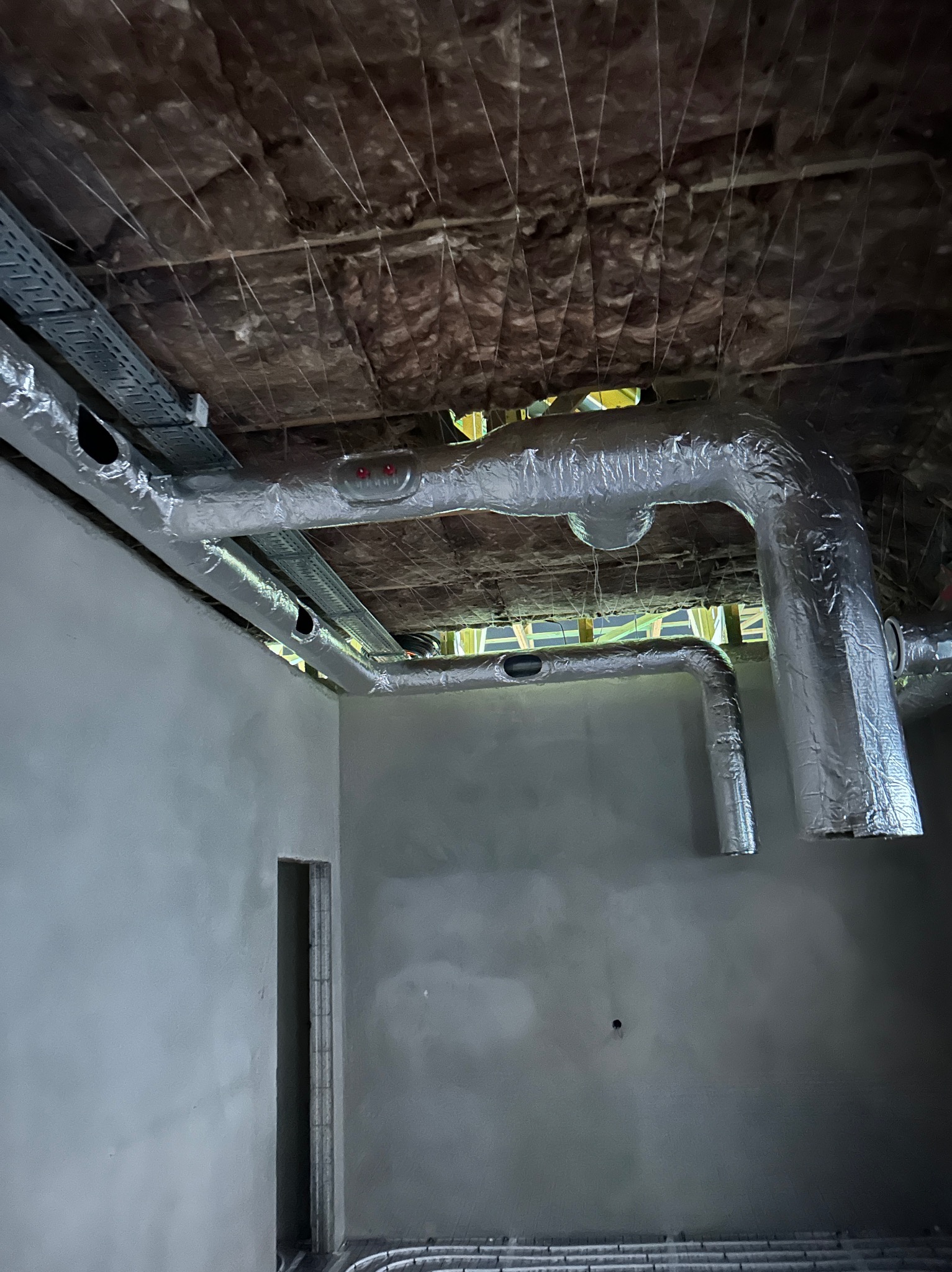 Szczegółowe ujęcie instalacji wentylacyjnej z aluminiową izolacją w surowym wnętrzu budynku w trakcie budowy, widoczne kable i izolacja stropu.