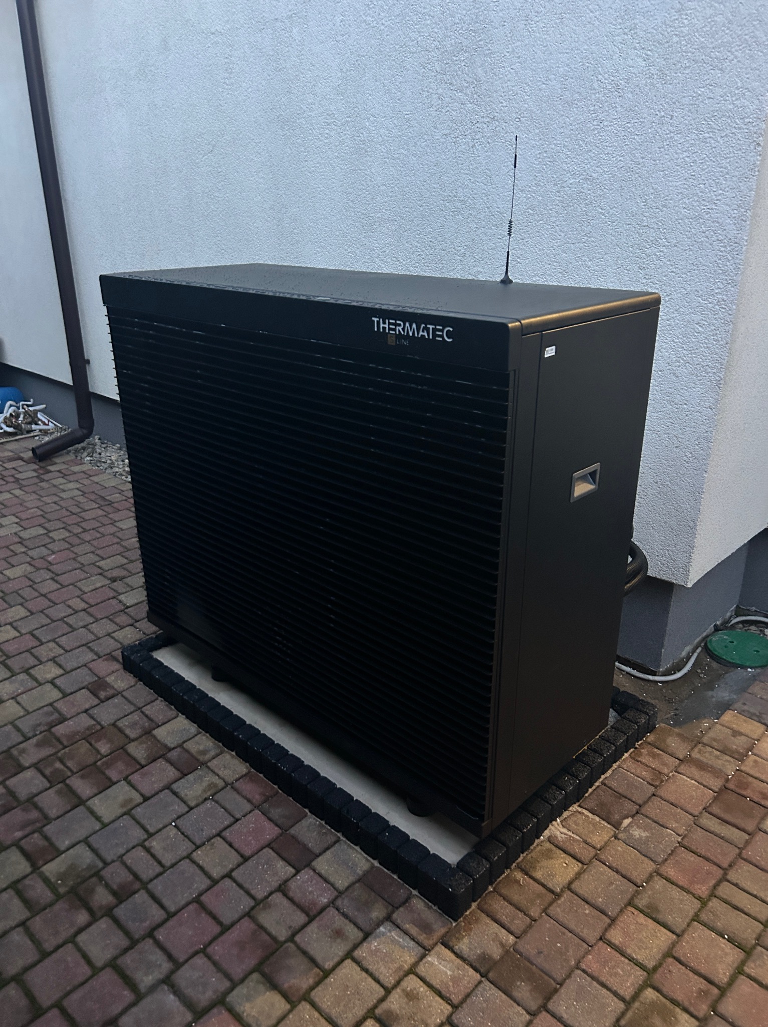 Czarna pompa ciepła marki Thermatec Line zainstalowana na zewnątrz budynku, widoczna antena na górze urządzenia, kostka brukowa wokół.