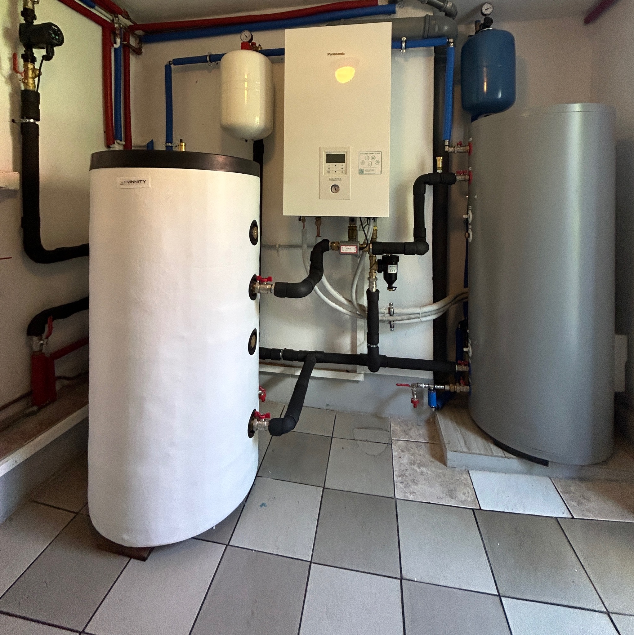 Instalacja kotła gazowego Panasonic z dwoma zbiornikami, białym Trinity i szarym, rurami w kolorach czerwonym i niebieskim, na podłodze z kwadratowych płytek.