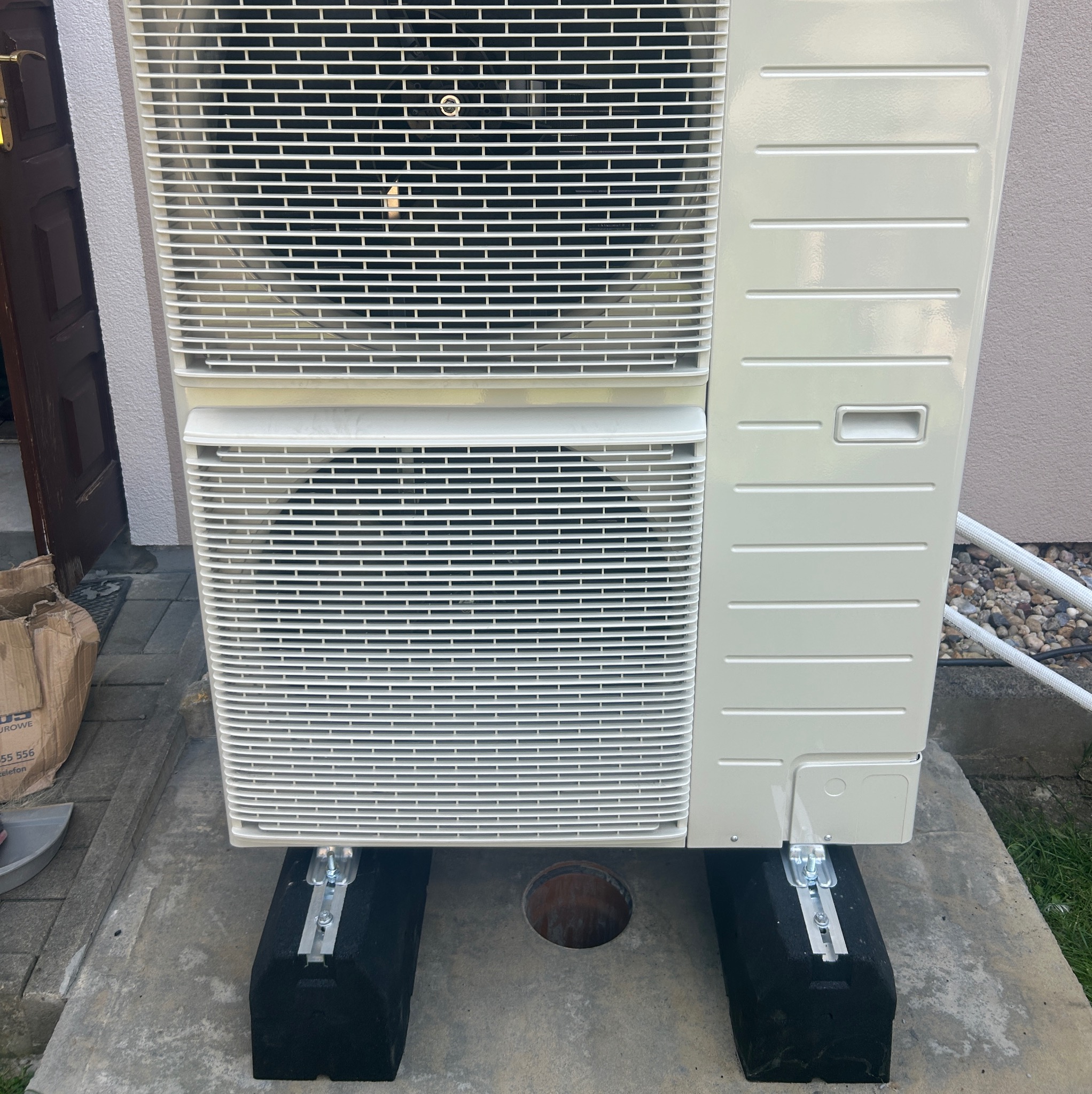 Zewnętrzna jednostka klimatyzacji umieszczona na czarnych podkładkach z aluminiowymi szynami, widoczny otwór w betonowej płycie, rury instalacyjne w tle.