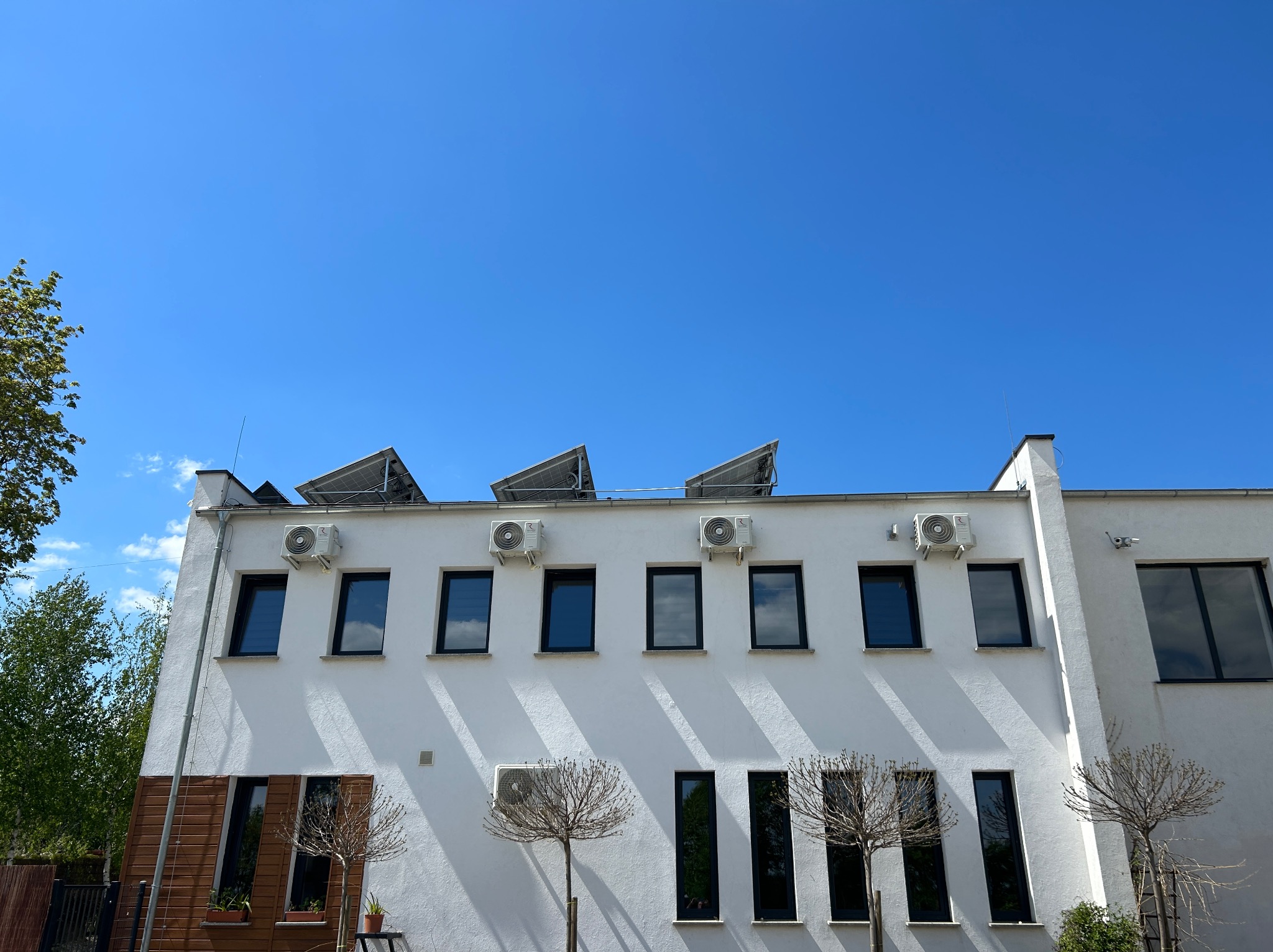 Biały budynek z rzędem okien i klimatyzatorów zamontowanych na elewacji pod dachem, z panelami słonecznymi na dachu, na tle błękitnego nieba.