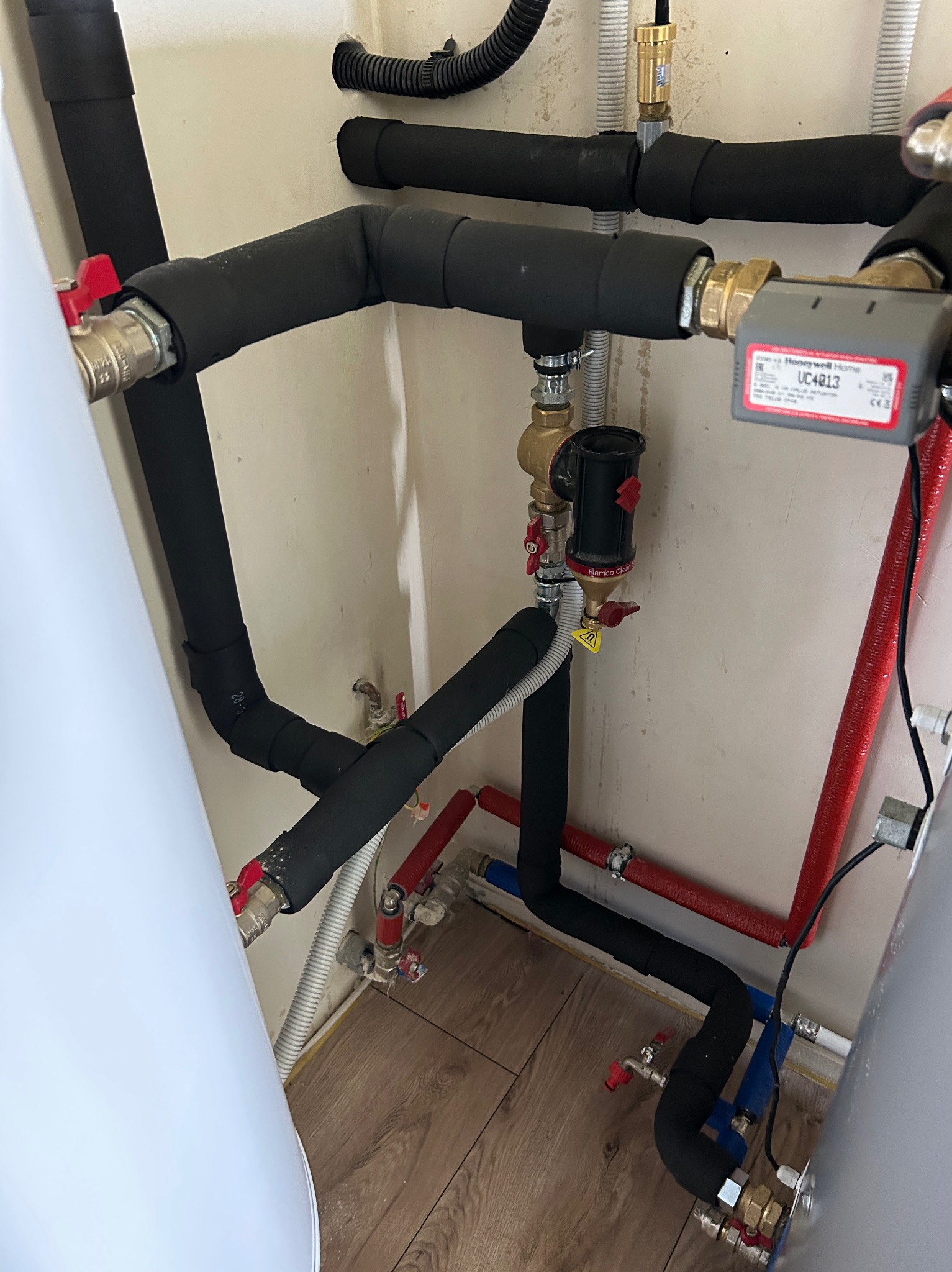 Instalacja grzewcza z czarnymi izolowanymi rurami, czerwonymi rurami, zaworami i filtrem Flamco Clean Smart zamontowana w pomieszczeniu z panelami podłogowymi.