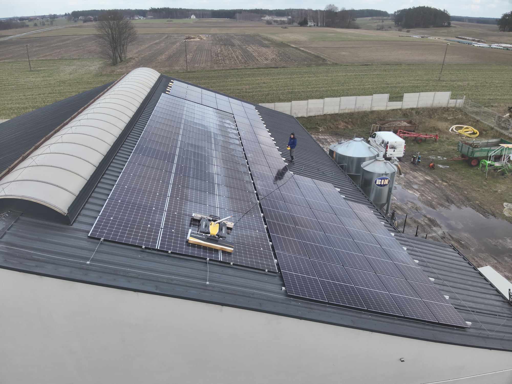 Czyszczenie paneli fotowoltaicznych na dachu budynku gospodarczego z użyciem automatycznego robota czyszczącego; w tle widoczne silosy i pole.