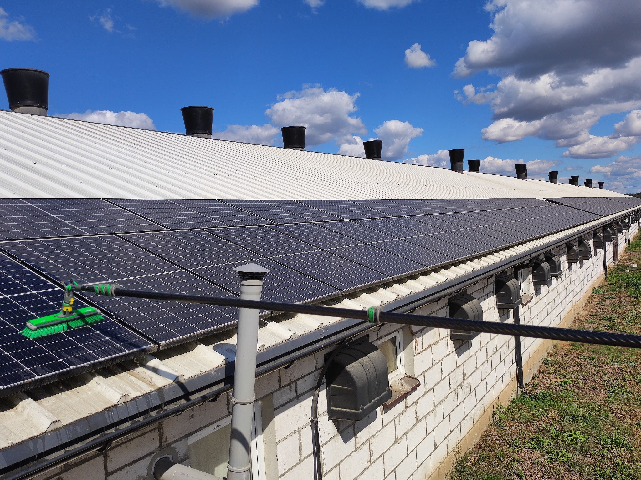 Czyszczenie paneli fotowoltaicznych na dachu budynku gospodarczego za pomocą szczotki na wysięgniku; biała cegła, wentylacja i błękitne niebo w tle.