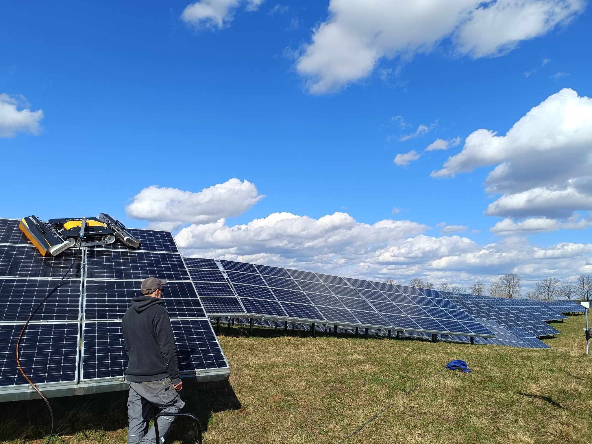 Czyszczenie paneli fotowoltaicznych za pomocą robota czyszczącego, pracownik nadzoruje proces, w tle rozległa farma paneli i błękitne niebo z obłokami.