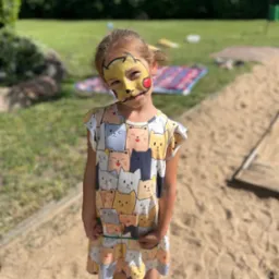 Dziewczynka z pomalowaną twarzą w motywie Pikachu stoi na piasku, w tle trawnik i drzewa.