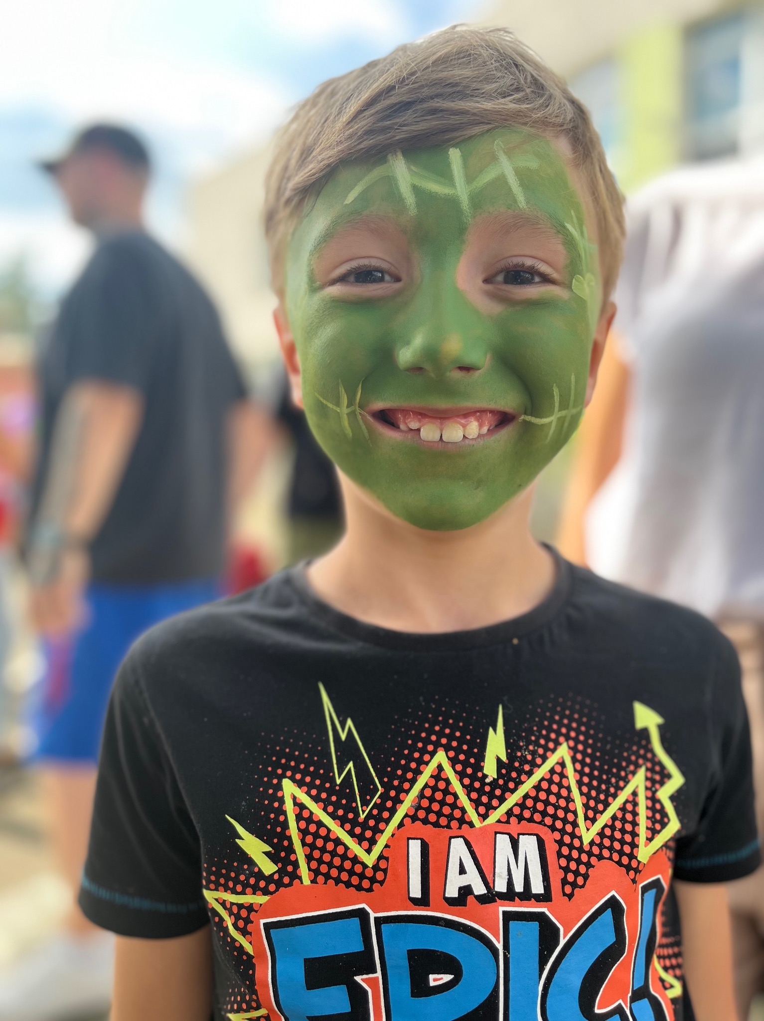 Uśmiechnięty chłopiec z zieloną farbą na twarzy, stylizowany na potwora Frankensteina, w czarnej koszulce z kolorowym nadrukiem, na rozmytym tle.