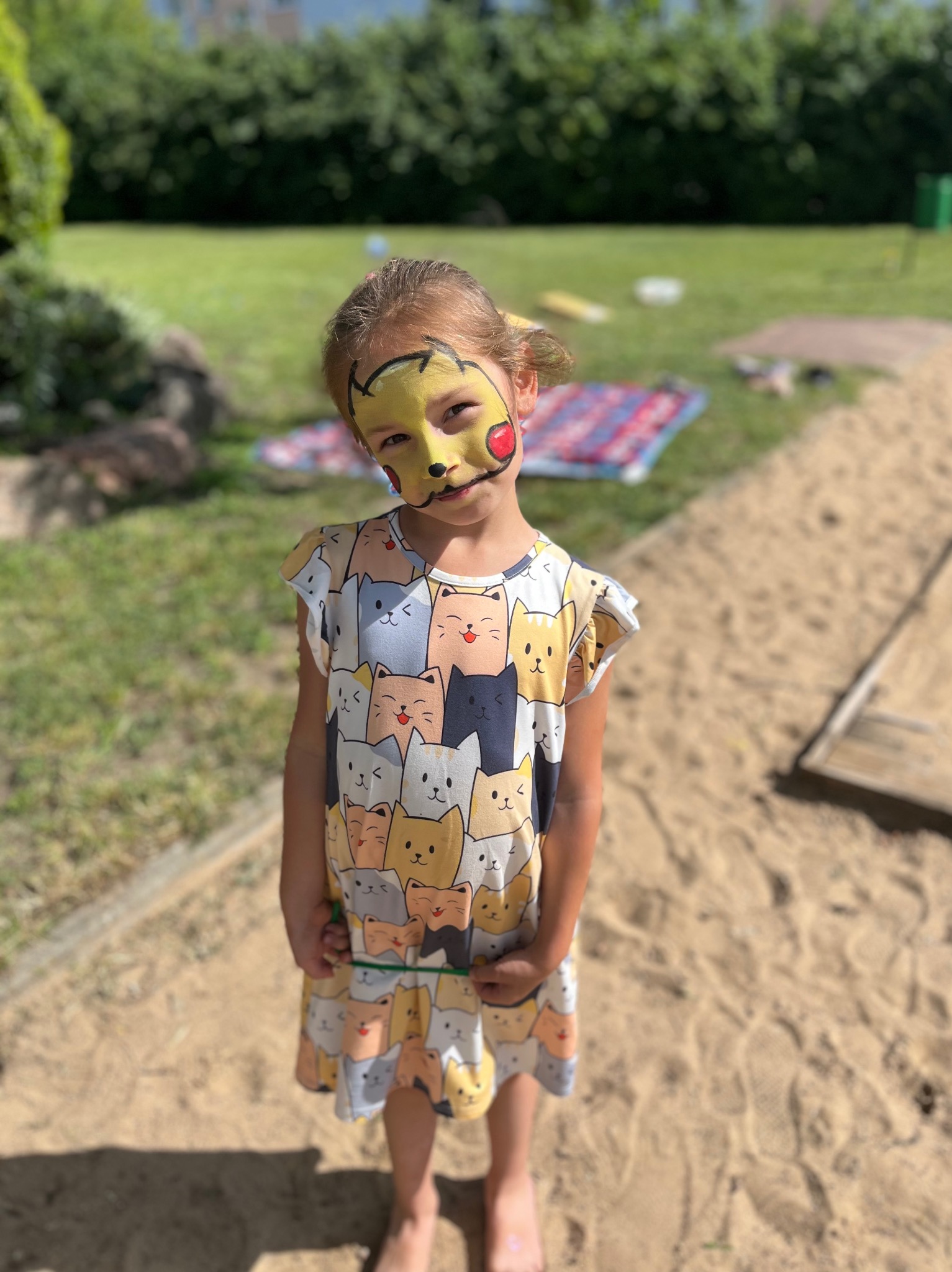 Dziewczynka z pomalowaną twarzą w motywie Pikachu stoi na piasku, w tle trawnik i drzewa.
