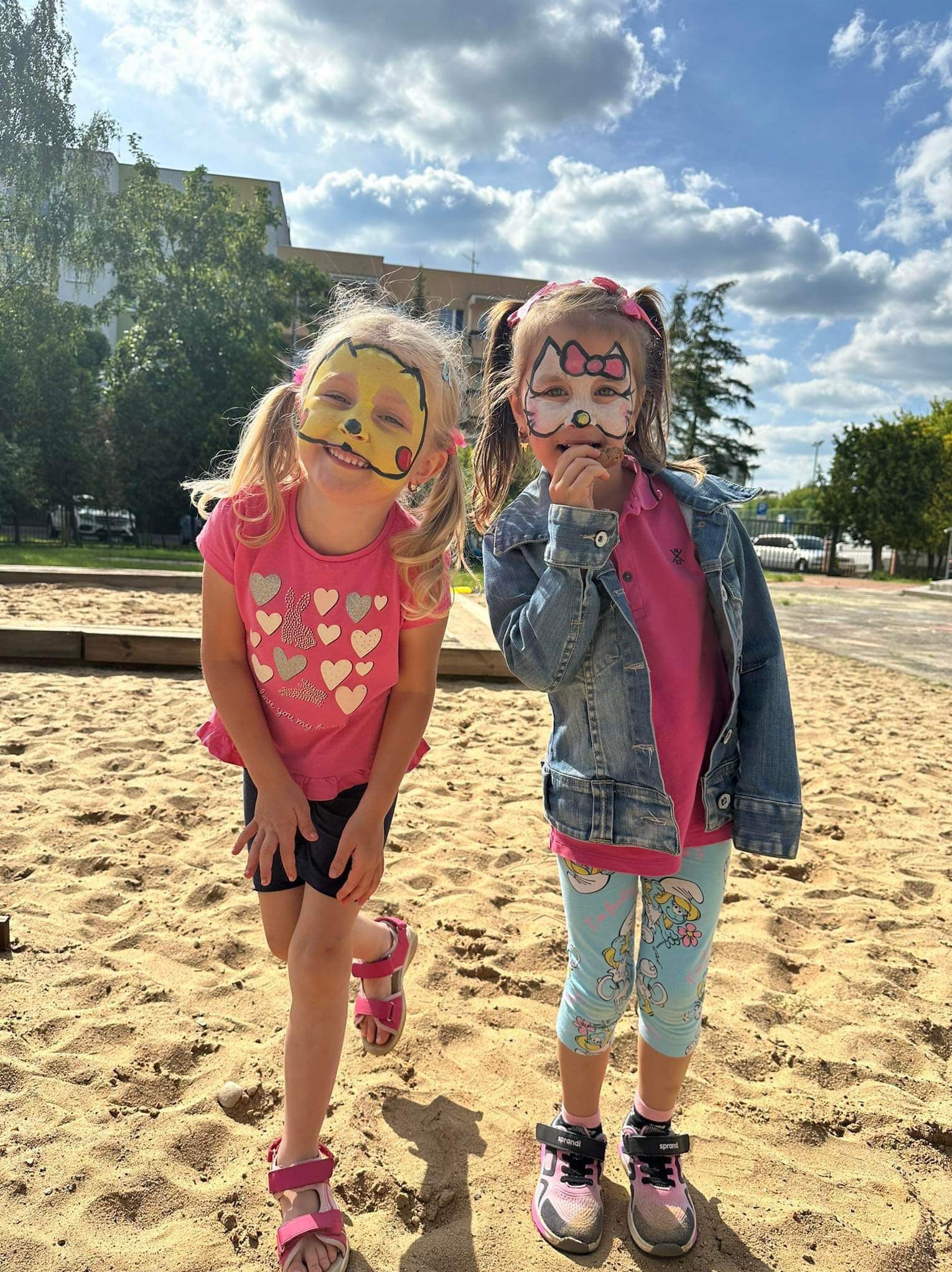 Dwie uśmiechnięte dziewczynki z pomalowanymi twarzami (motyw Pikachu i Hello Kitty) stoją w piaskownicy na tle drzew i błękitnego nieba z chmurami.