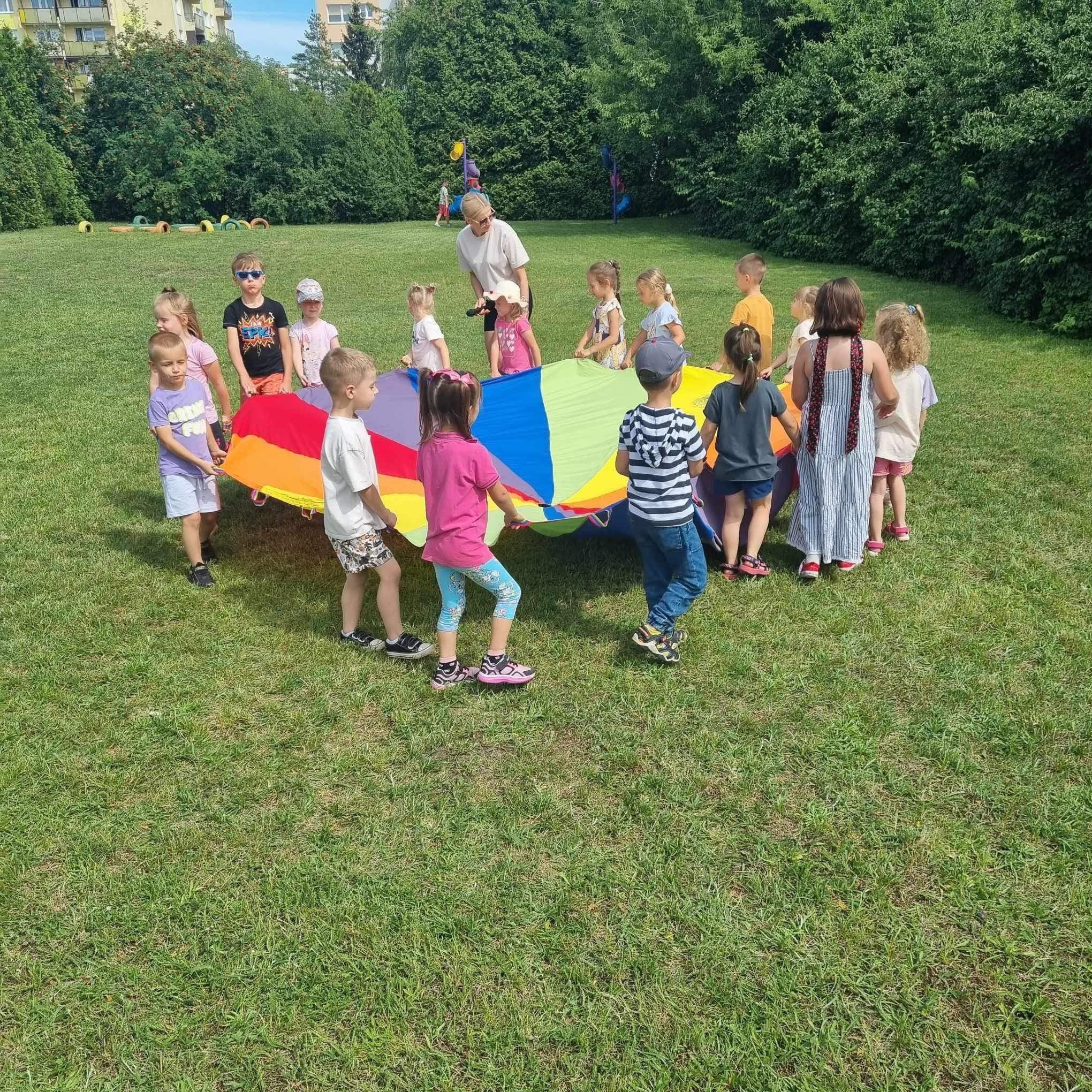 Grupa dzieci bawi się na trawie kolorową chustą animacyjną, nadzorowana przez dorosłą osobę, w tle budynek mieszkalny i drzewa.