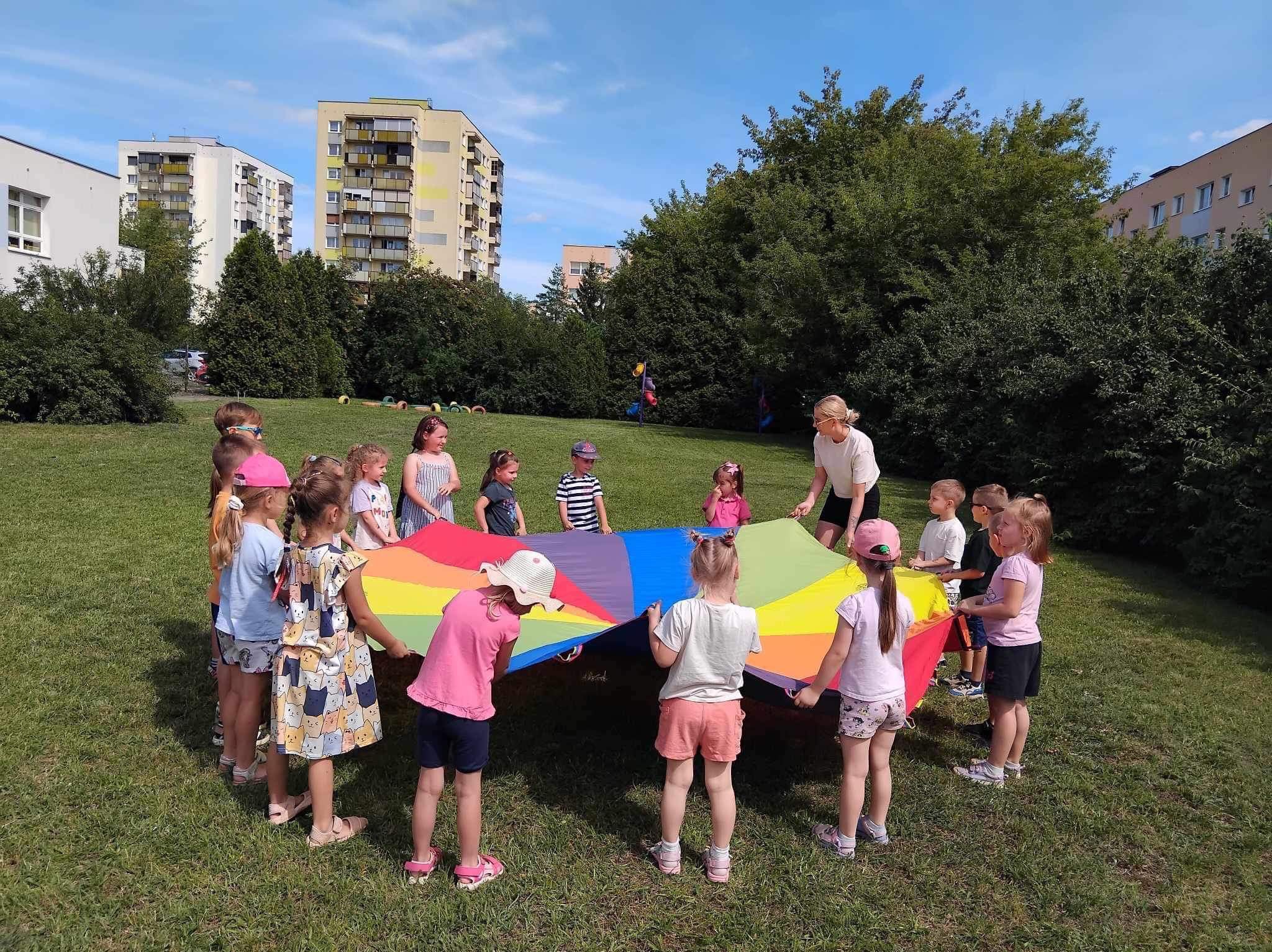 Grupa dzieci trzymająca kolorową chustę animacyjną na trawie, w tle zabudowa miejska i drzewa.