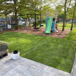 New Eden Usługi Ogrodnicze Mateusz Kazimierski - Świeżo ułożona trawa z rolki na placu zabaw, z widocznym stosem rolek trawy i wiaderkiem. W tle huśtawka i zjeżdżalnia. Ciekawy efekt wizualny.