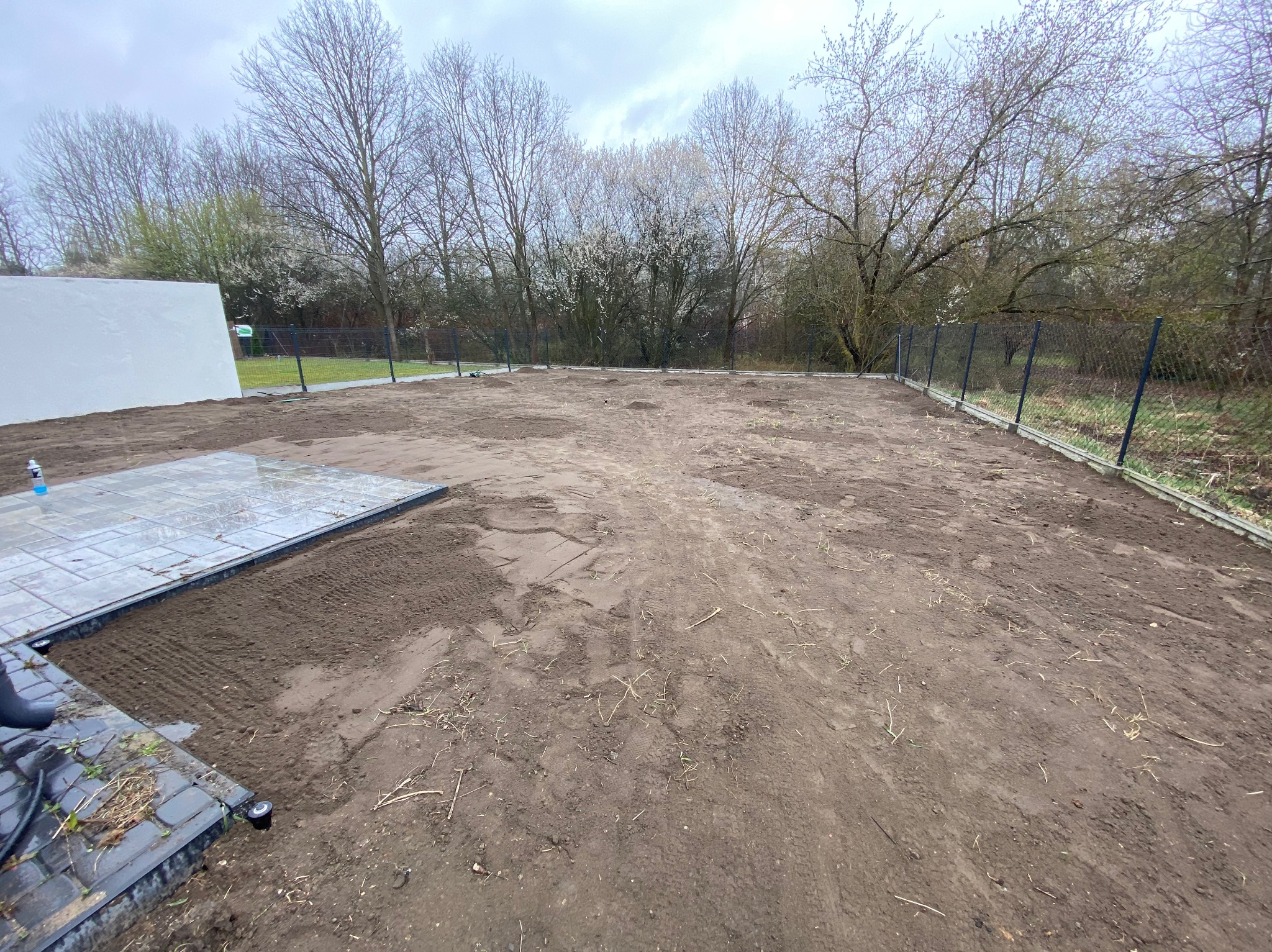 Przygotowany teren pod trawnik z rolki. Widoczne świeżo rozłożone płyty chodnikowe, ogrodzenie, drzewa w tle i pochmurne niebo. Wyrównana ziemia czeka na ułożenie trawy.