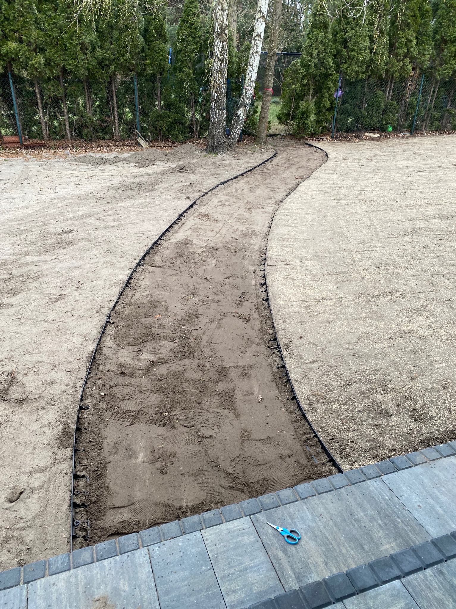 Wyrównany teren pod ścieżkę w ogrodzie z obrzeżami z tworzywa, widoczne narzędzie (nożyczki) na kostce brukowej. W tle drzewa iglaste i brzoza.