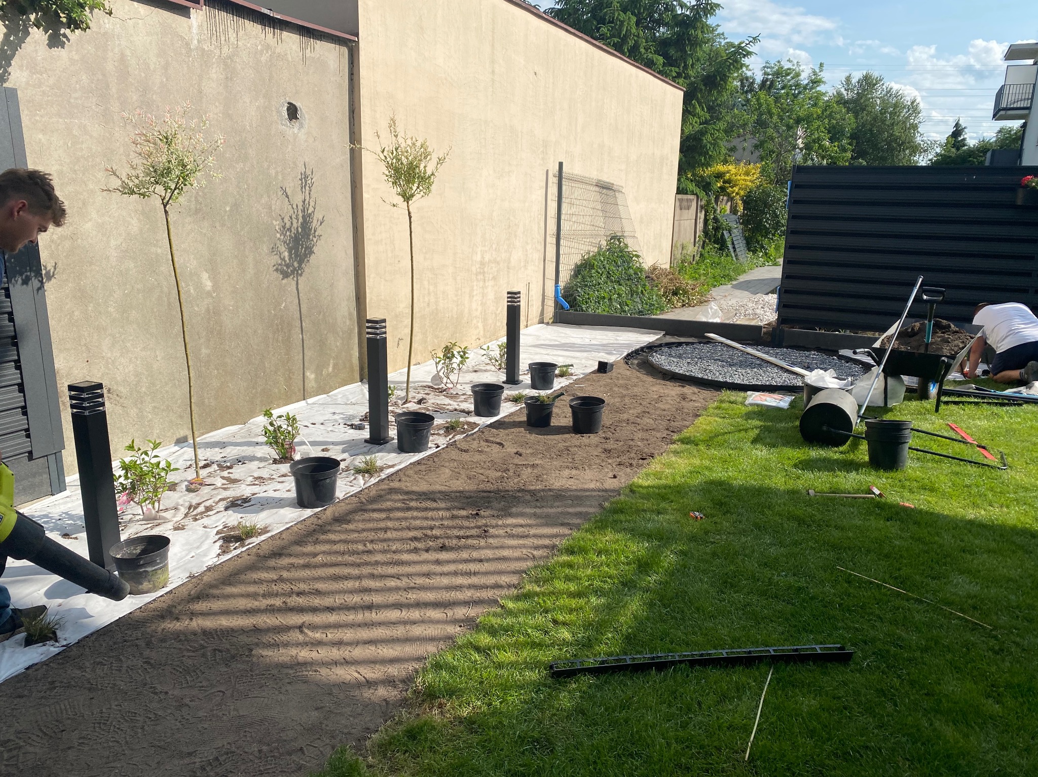 Prace ogrodowe w toku: przygotowanie podłoża, sadzenie drzewek i montaż oświetlenia wzdłuż ścieżki na posesji. Widoczne narzędzia i elementy dekoracyjne.