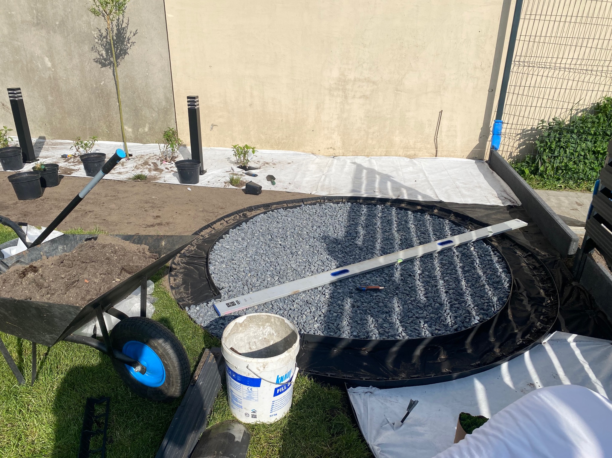 Okrągła rabata z szarym żwirem w ogrodzie. Widoczna taczka z ziemią, poziomica, wiadro i sadzonki. Etap tworzenia ogrodu, z bliska, w słoneczny dzień.