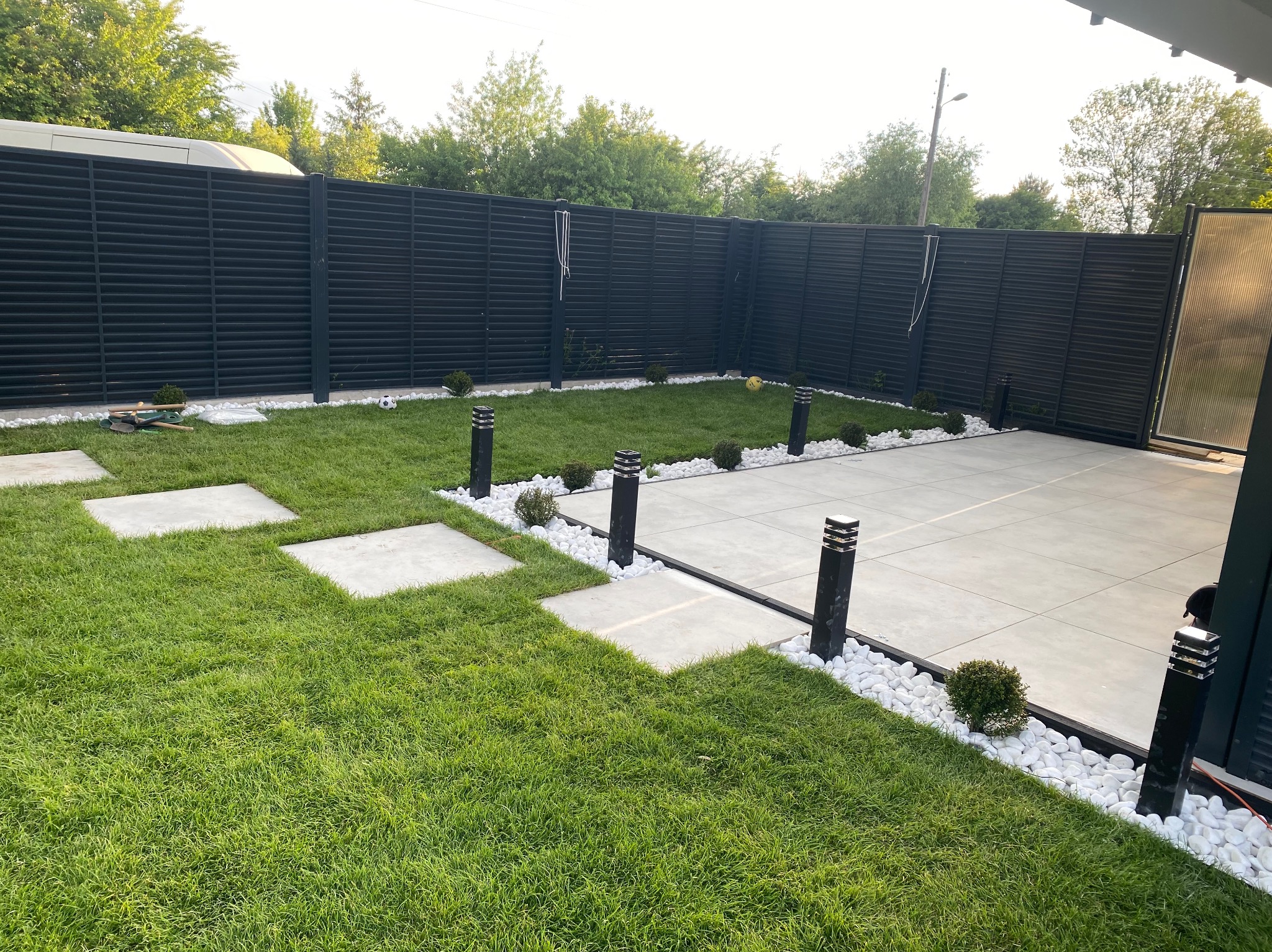 Nowoczesny ogród z trawnikiem, ścieżką z płyt, białymi kamieniami i czarnym ogrodzeniem. Oświetlenie ogrodowe wzdłuż ścieżki. Minimalistyczny design.