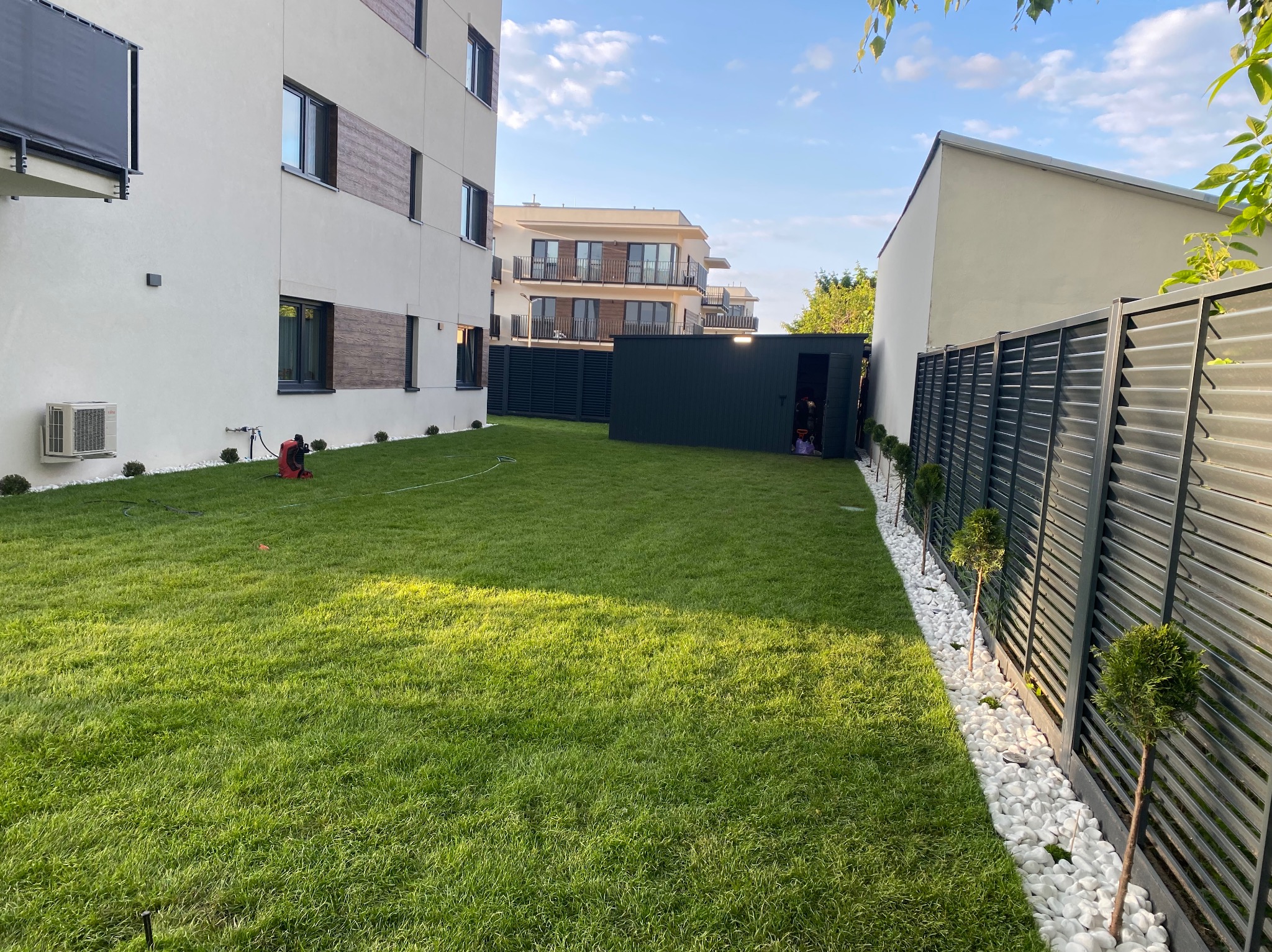 Zadbany trawnik z rolki przed nowoczesnym budynkiem, z widocznym systemem nawadniania i ogrodzeniem z białymi kamieniami. Estetyczna przestrzeń przydomowa.