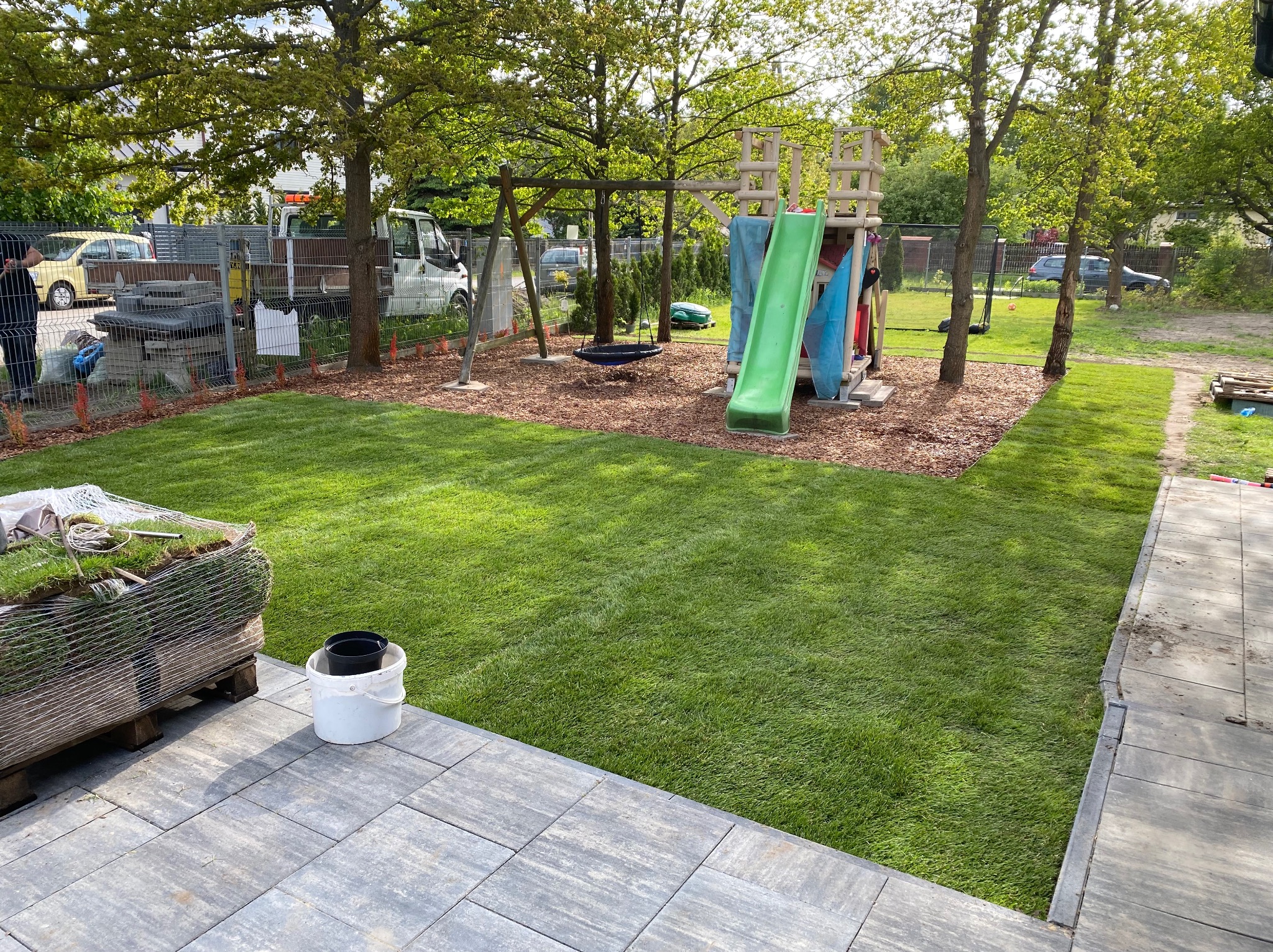 Świeżo ułożona trawa z rolki na placu zabaw, z widocznym stosem rolek trawy i wiaderkiem. W tle huśtawka i zjeżdżalnia. Ciekawy efekt wizualny.