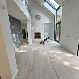 Przemo_Parkiety - Jasna, odnowiona podłoga parkietowa w przestronnym salonie z wysokim sufitem, widoczna maszyna do cyklinowania i długi kabel zasilający na podłodze.