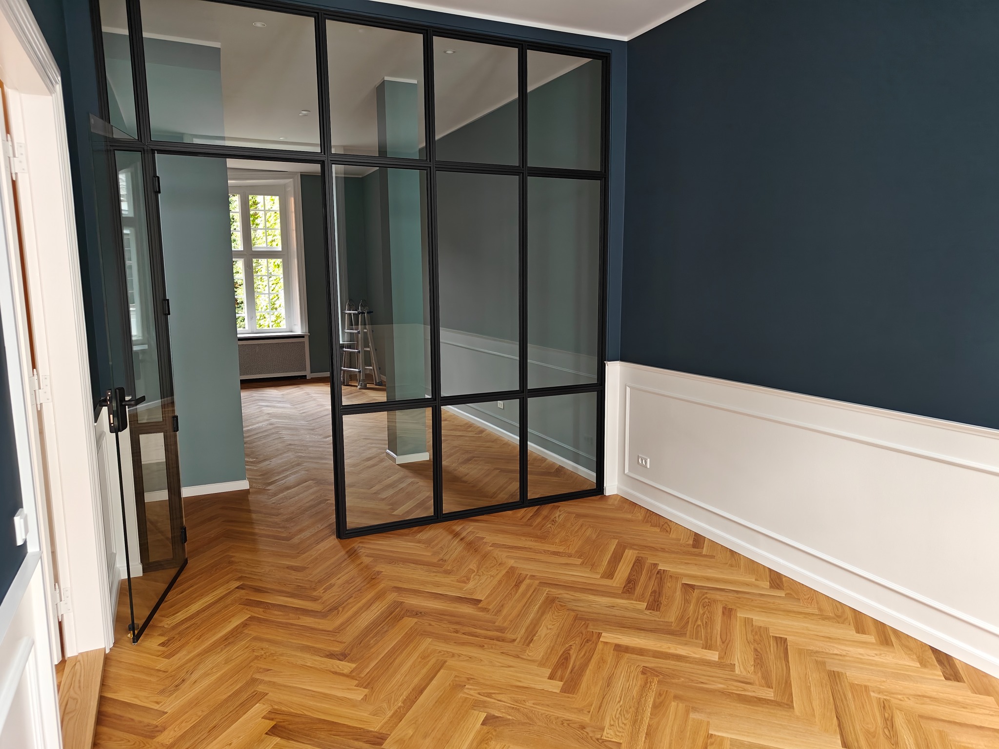 Parkiet ułożony w jodełkę francuską w przestronnym pokoju z granatową ścianą i białą boazerią, widoczne metalowe drzwi z szybami i drabina w tle.