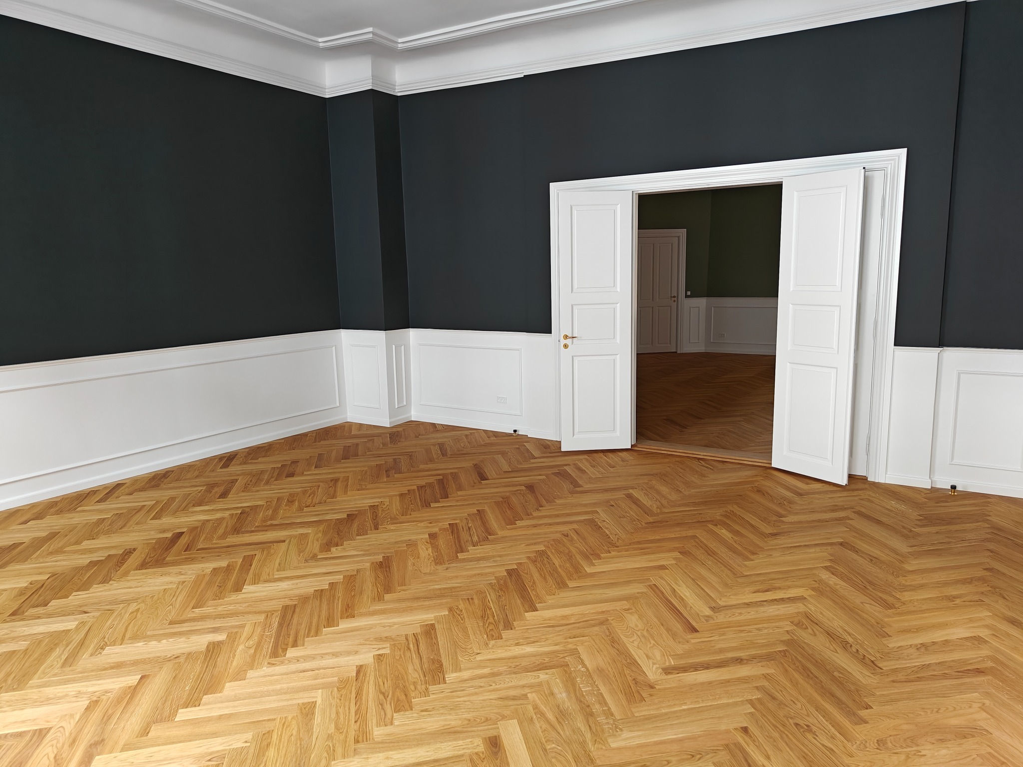 Jasny parkiet ułożony we wzór jodełki francuskiej w przestronnym, eleganckim pokoju z ciemnymi ścianami i białymi panelami ściennymi oraz otwartymi, białymi drzwiami do sąsiedniego pomieszczenia.