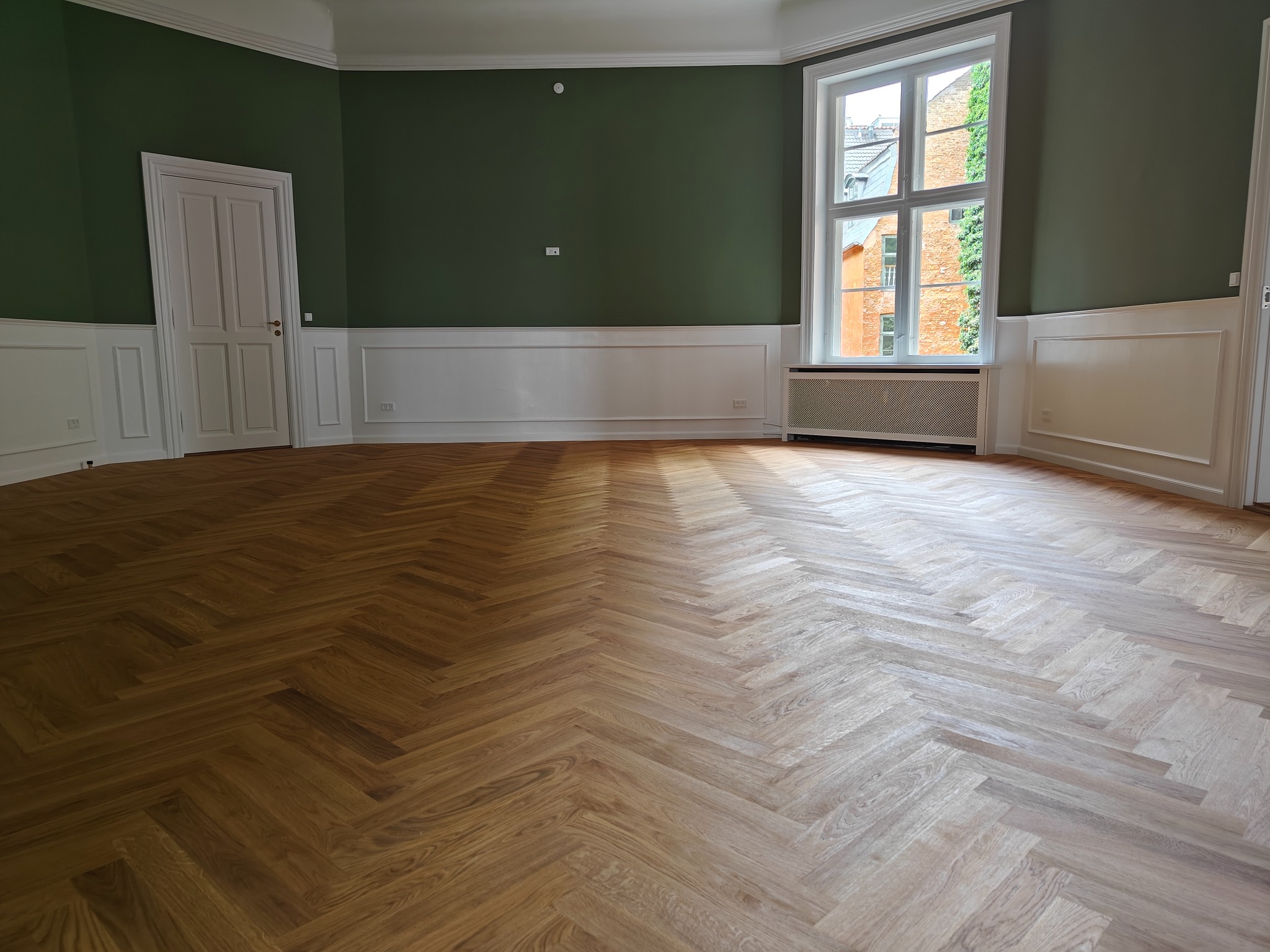Parkiet ułożony w jodełkę francuską w pustym, odnowionym pokoju z zielonymi ścianami i białymi panelami ściennymi, widok na budynki przez okno.