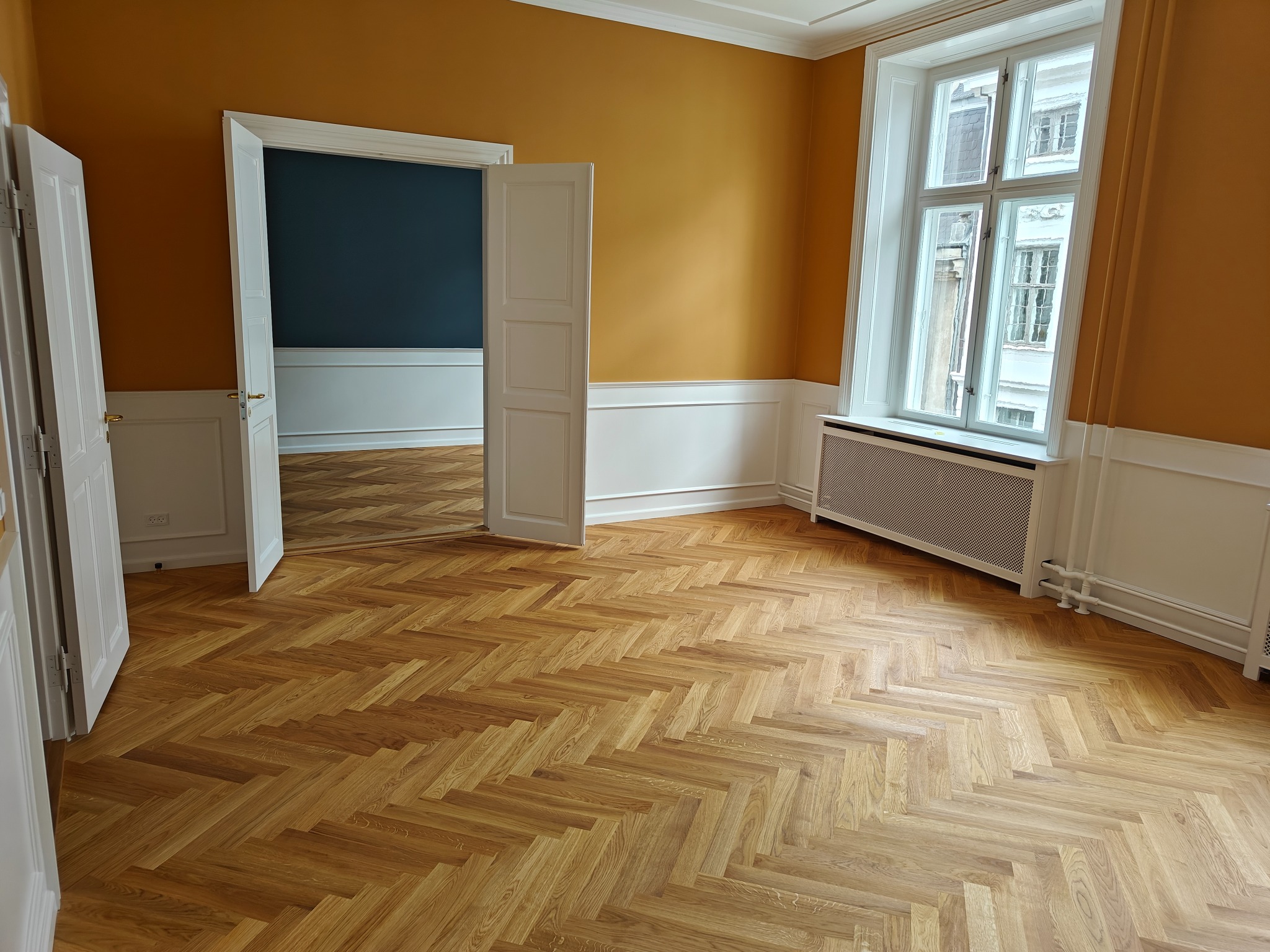 Jasny parkiet ułożony w jodełkę w przestronnym pokoju z pomarańczowymi ścianami i białymi panelami, widok na sąsiedni pokój z granatową ścianą przez otwarte białe drzwi.