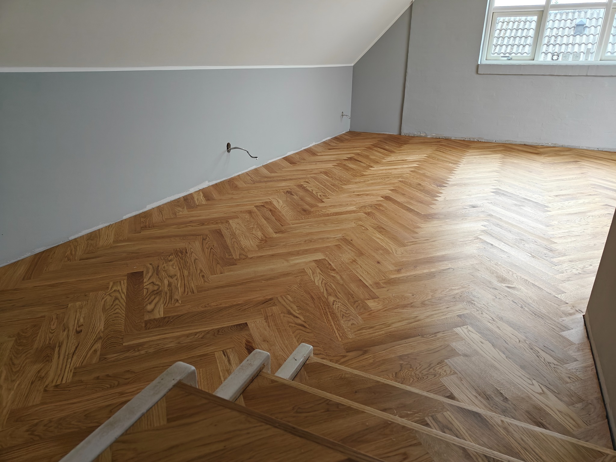 Nowo ułożony parkiet w jodełkę w poddaszowym pokoju, widok z góry schodów, szare ściany, widoczne gniazdko elektryczne z wystającym kablem.