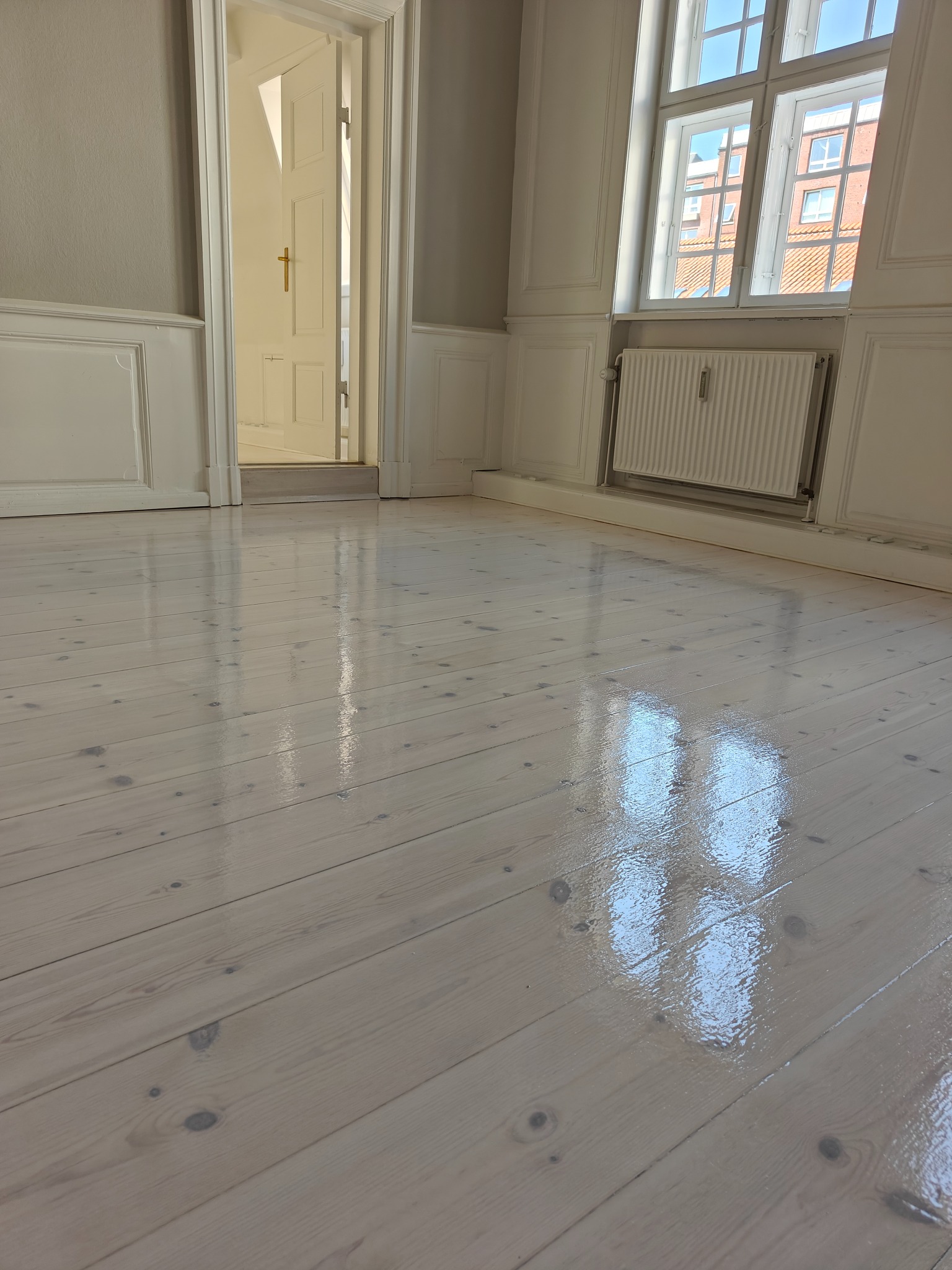 Bielona, lakierowana podłoga z desek w jasnym pokoju z białymi panelami ściennymi, widoczne okno i otwarte drzwi do kolejnego pomieszczenia.