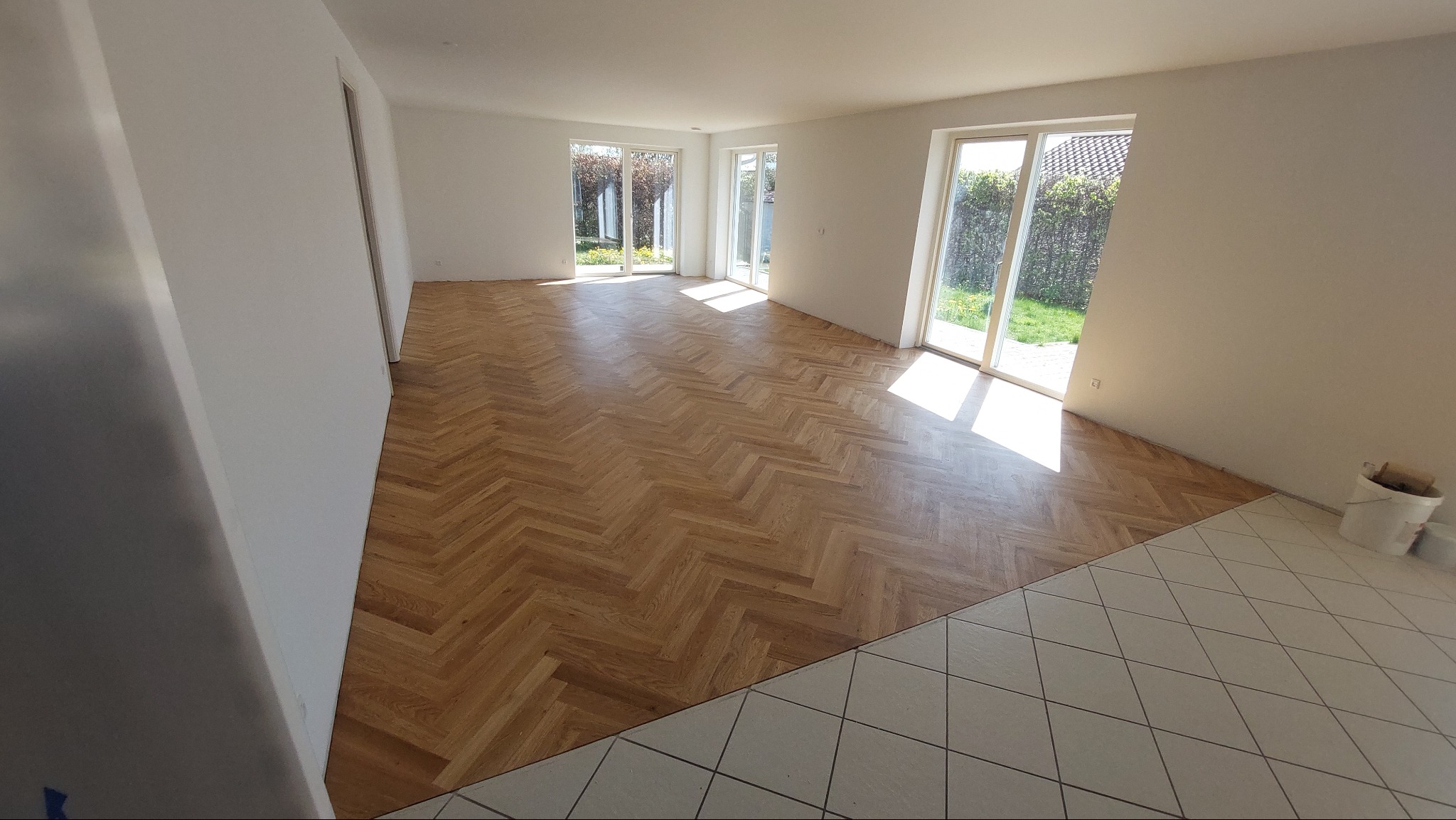 Nowo ułożony parkiet w jodełkę w przestronnym, jasnym salonie z widokiem na ogród przez przeszklone drzwi balkonowe; fragment podłogi wykończony białymi płytkami.