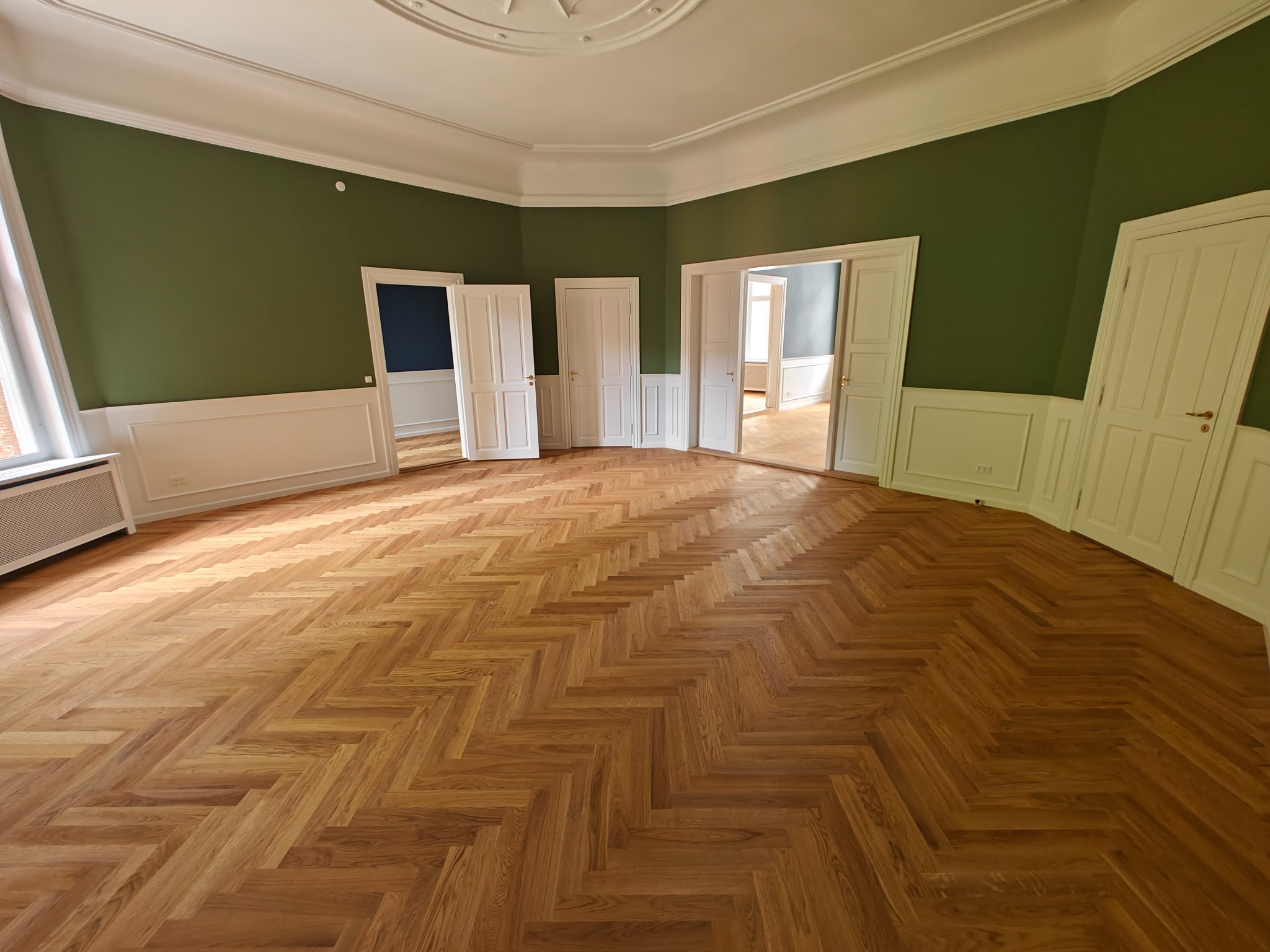 Wnętrze przestronnego pokoju z parkietem ułożonym w jodełkę, zielonymi ścianami z białymi panelami i kilkoma parami białych drzwi.