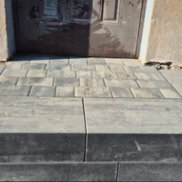 Schody wejściowe wyłożone kostką brukową w odcieniach szarości, prowadzące do ciemnych drzwi. Widoczne fragmenty ściany budynku z różną fakturą tynku.