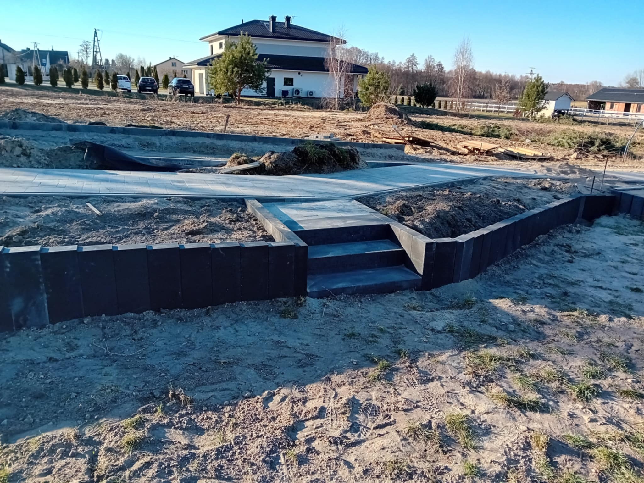 Nowo ułożona, szara kostka brukowa prowadzi do domu. W tle widoczny budynek, drzewa i zaparkowane samochody. Całość w słoneczny dzień.