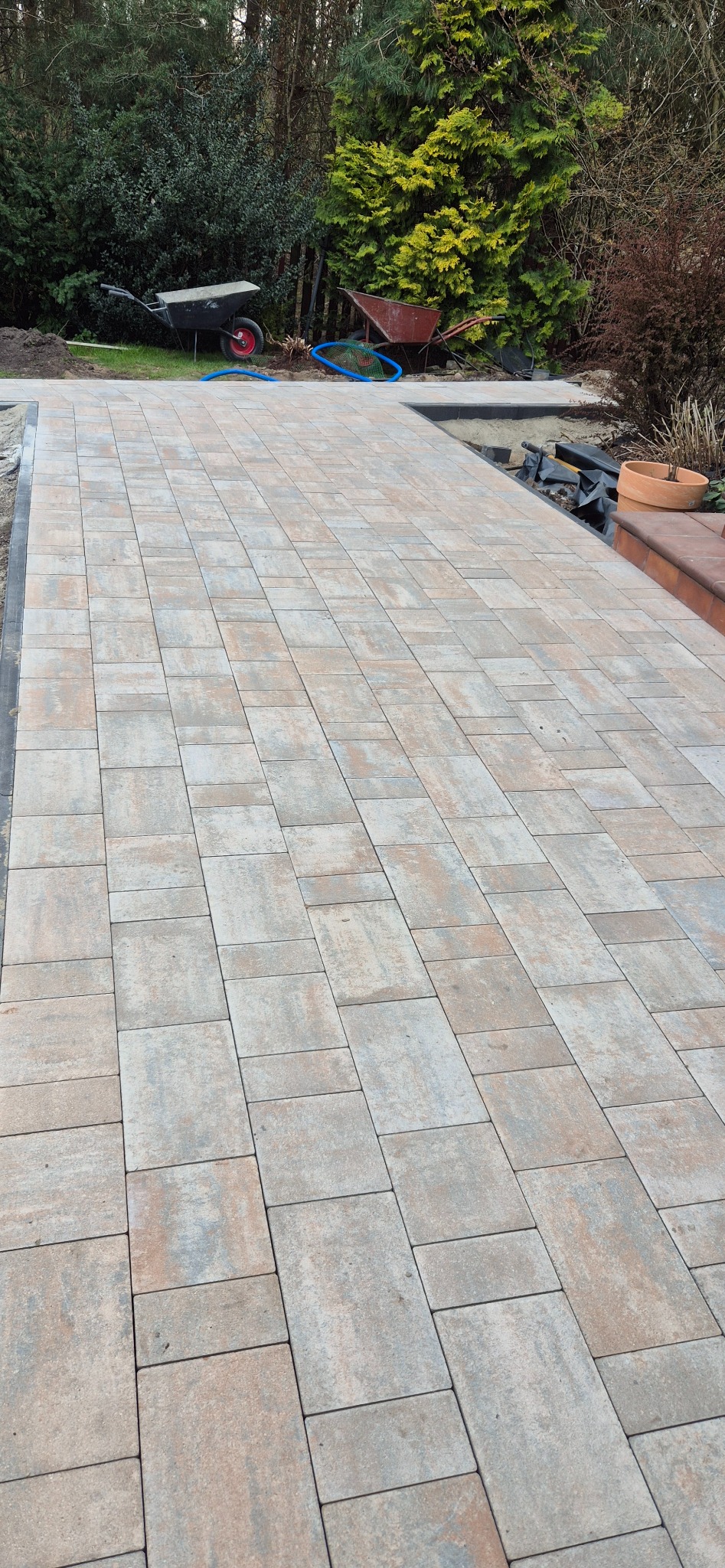 Ukończona ścieżka z beżowo-szarych kostek brukowych, ułożonych w regularny wzór. W tle widoczne narzędzia ogrodowe i roślinność.