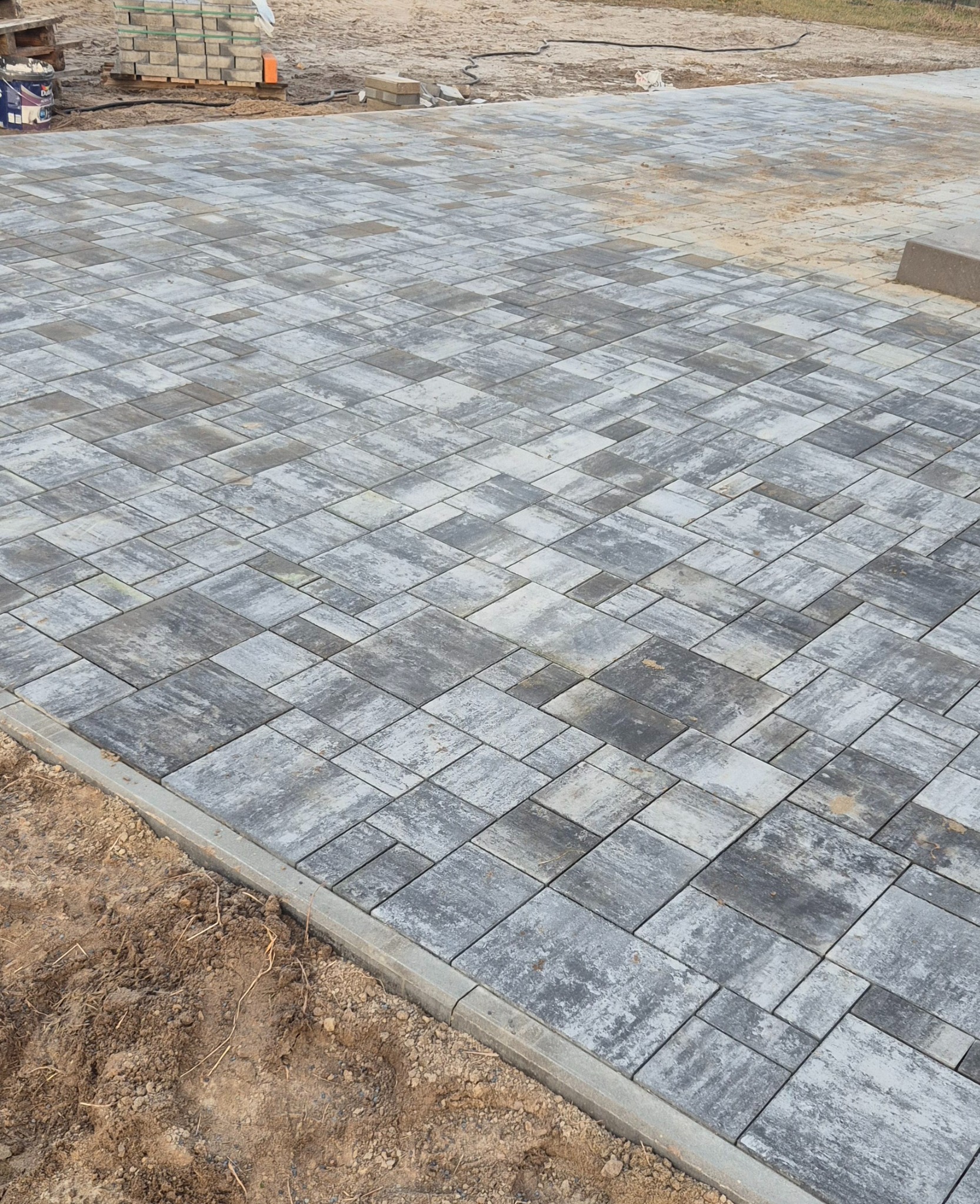 Ułożona, szara kostka brukowa o nieregularnym wzorze, obramowana betonowym krawężnikiem. W tle sterta kostki i piasek.