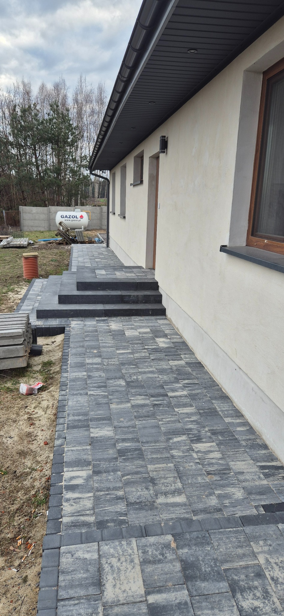 Ułożona kostka brukowa wzdłuż elewacji domu, prowadząca do schodów wejściowych. Widoczny fragment elewacji, okna i tło z drzewami. Szara kostka tworzy chodnik.