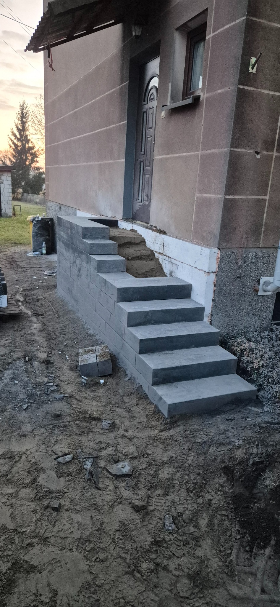 Nowe, szare schody wejściowe z kostki przy domu. Widoczne ślady prac ziemnych i częściowo odsłonięta podmurówka budynku.
